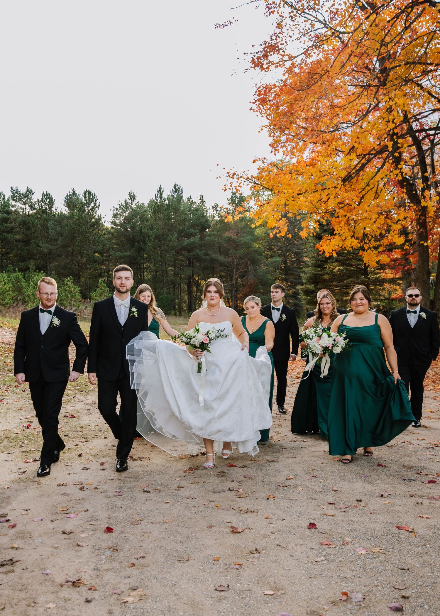 Wedding party walks together on sandy ground surrounded by autumn trees with vibrant orange foliage.