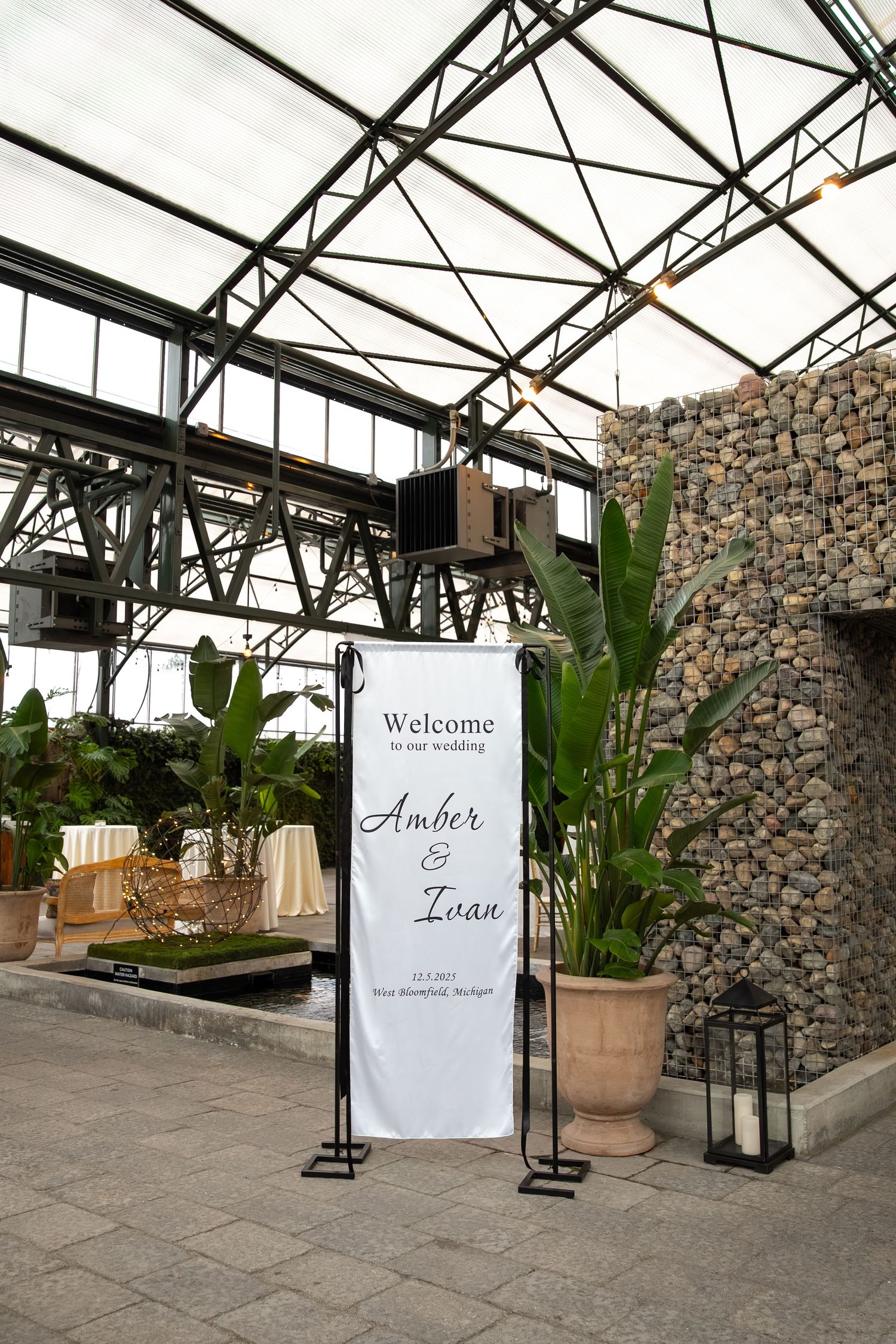 Welcome banner for Amber and Ivan wedding at a greenhouse venue in Bloomfield Michigan with tropical plants and stone wall.
