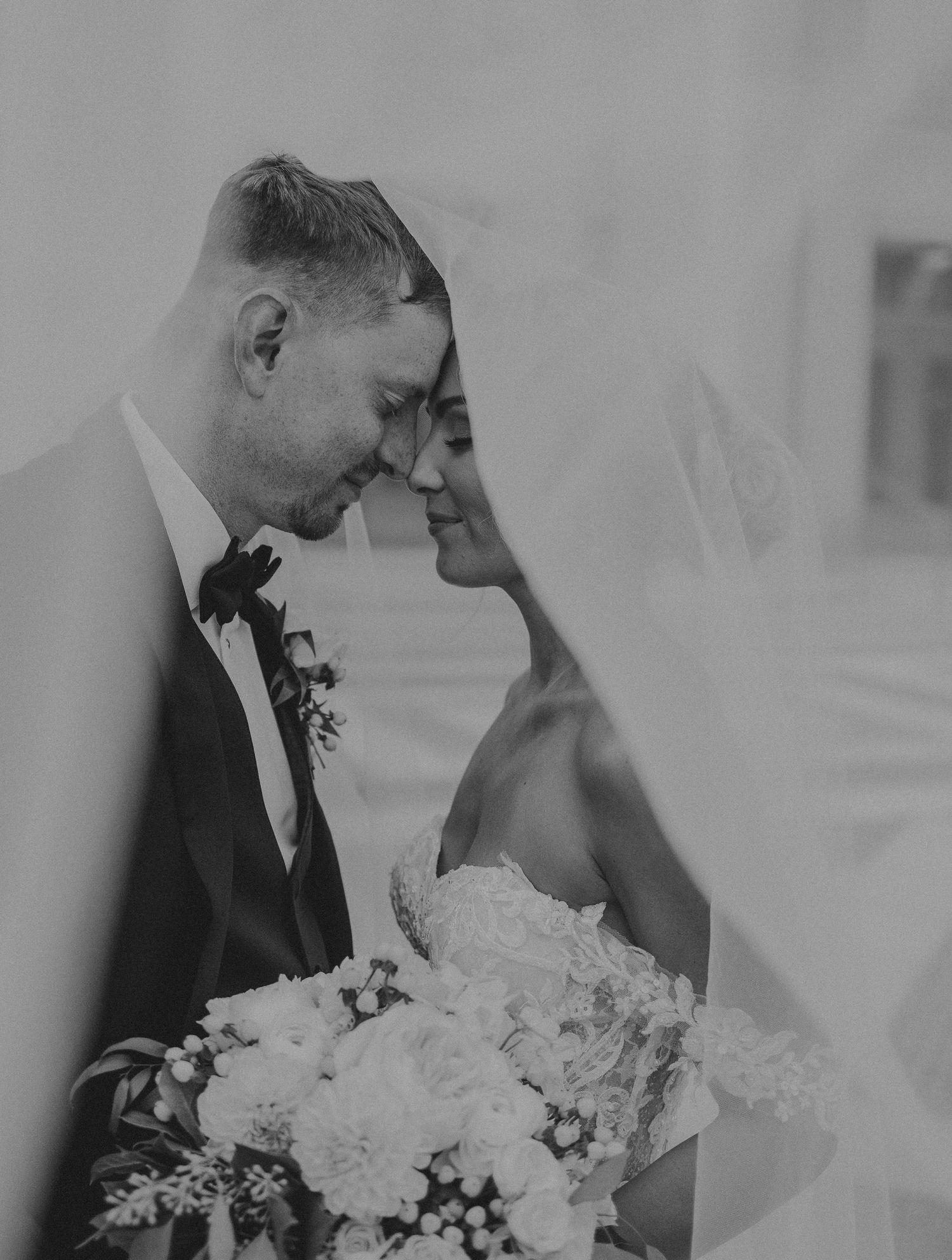 Black and white intimate wedding portrait showing a tender moment between newlyweds with white floral bouquet.