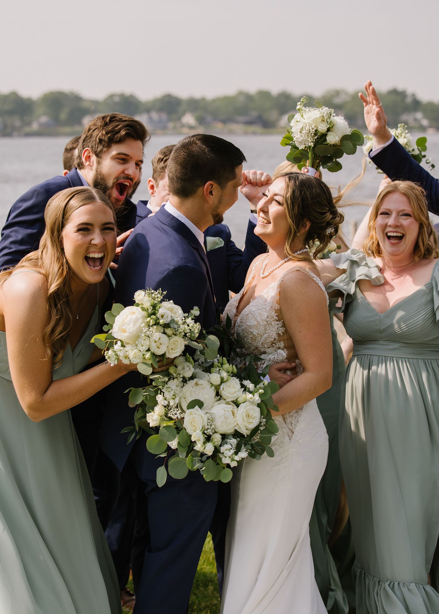 Wedding party celebrates joyfully together at outdoor waterfront venue in sage green and navy attire.