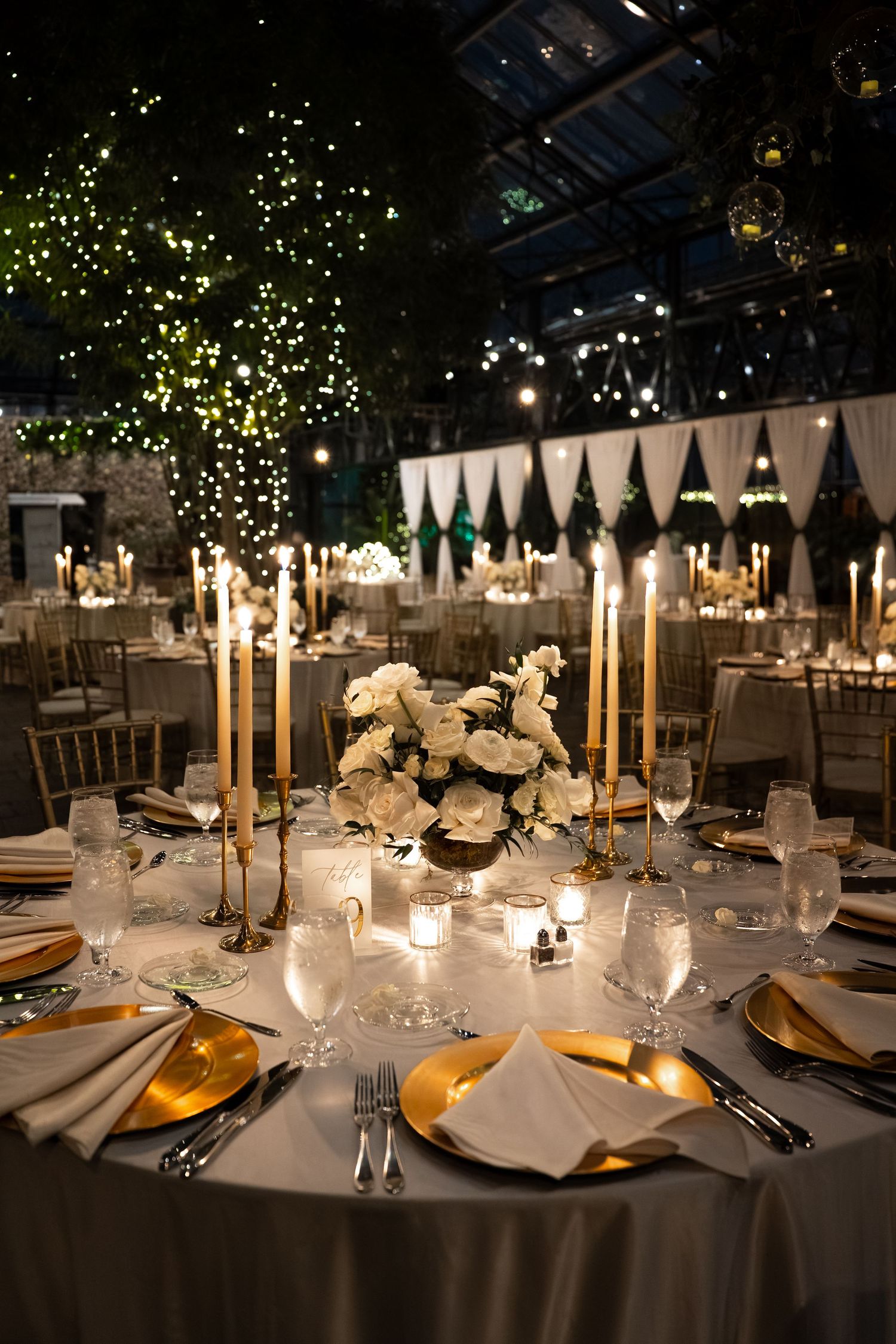 Elegant wedding reception table with gold candelabras, white roses, gold chargers, and glowing fairy lights in a greenhouse venue.