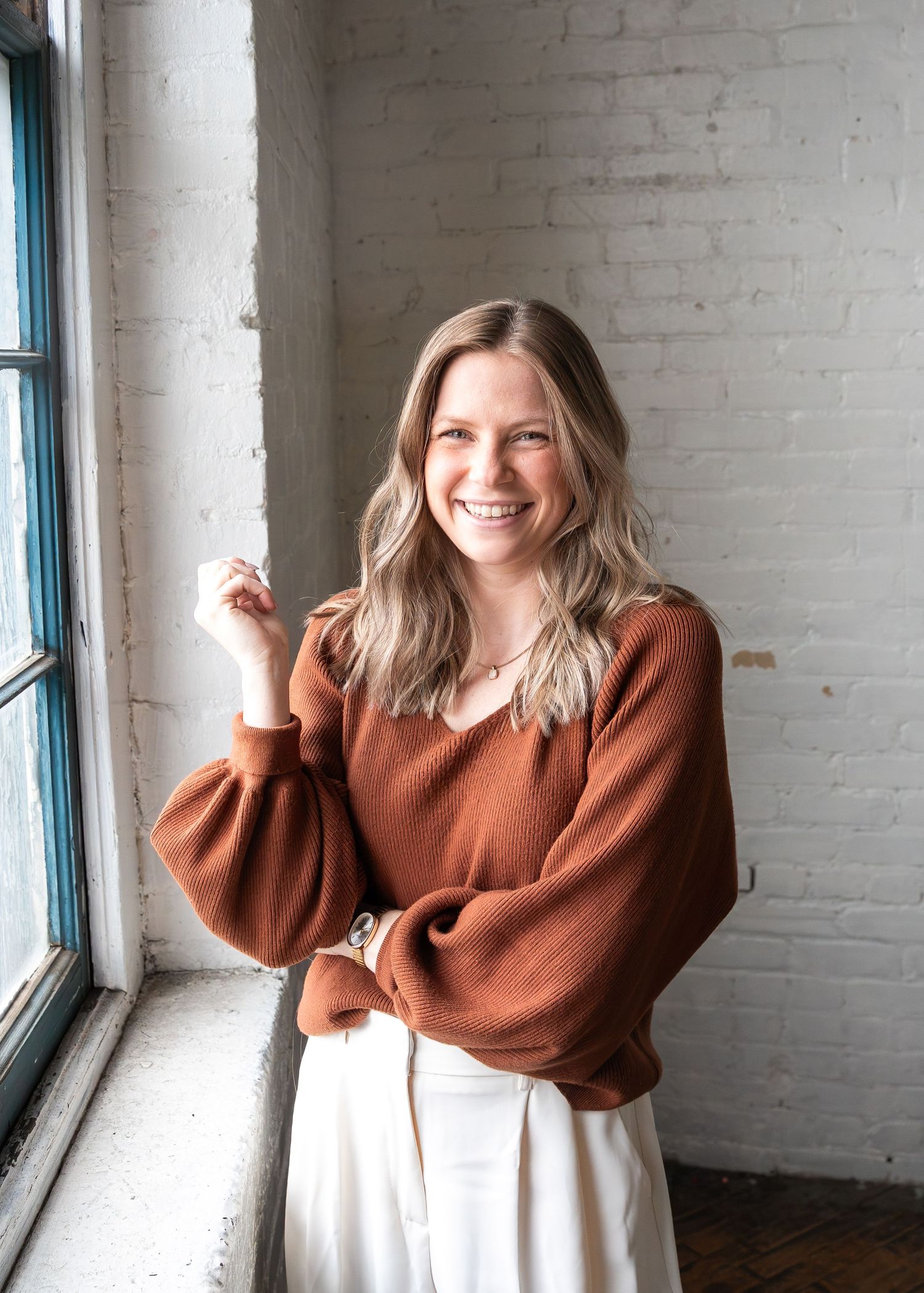 Smiling woman in rust orange sweater and white skirt poses by window in bright brick interior.