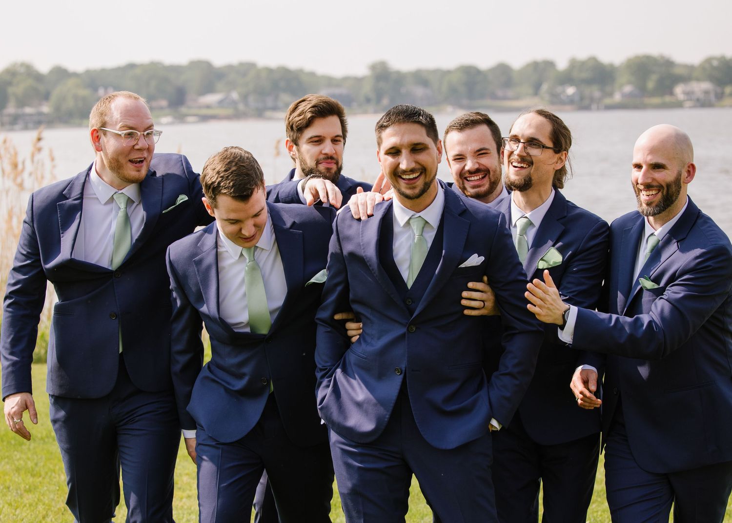Groomsmen in navy suits share a candid laughing moment together on a lawn by a lake.
