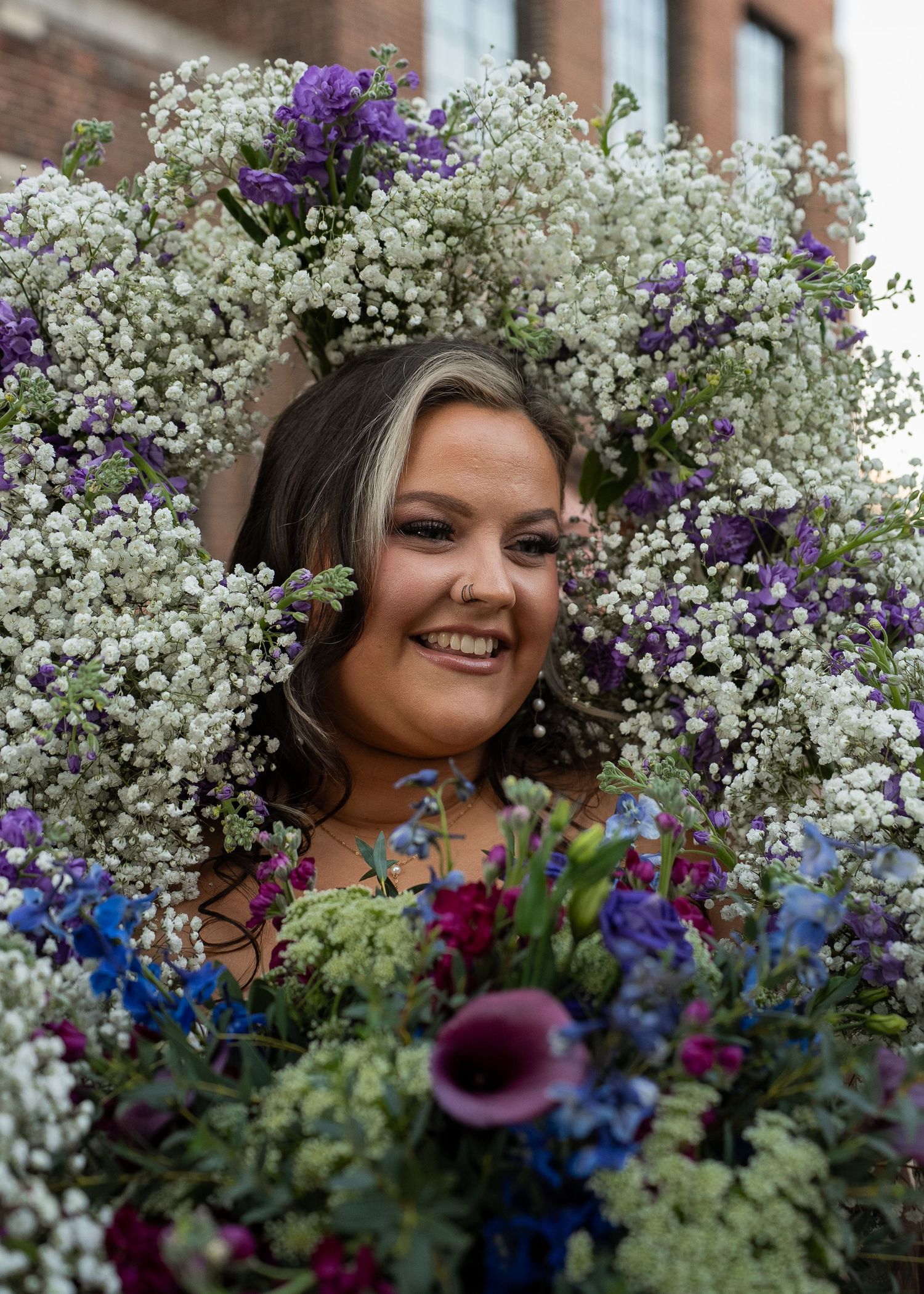 Smiling woman surrounded by large floral arrangement of white baby's breath, purple and blue flowers outdoors.