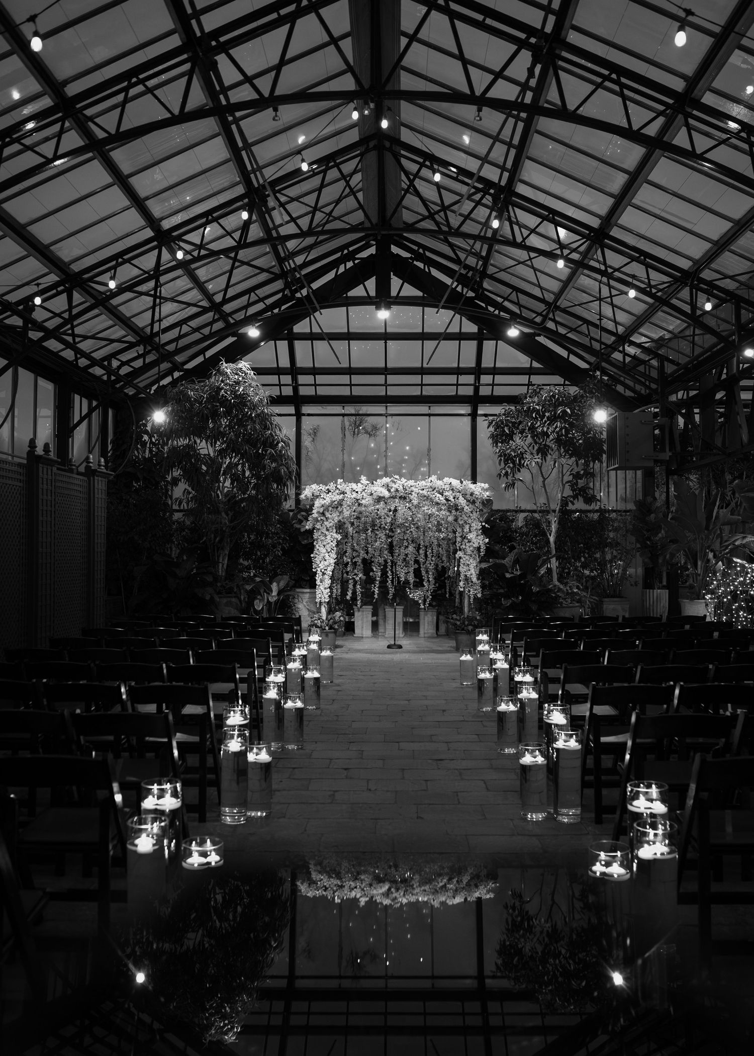 Black and white photo of an elegant indoor wedding ceremony venue with floral arch, candlelit aisle, and greenhouse glass ceiling.