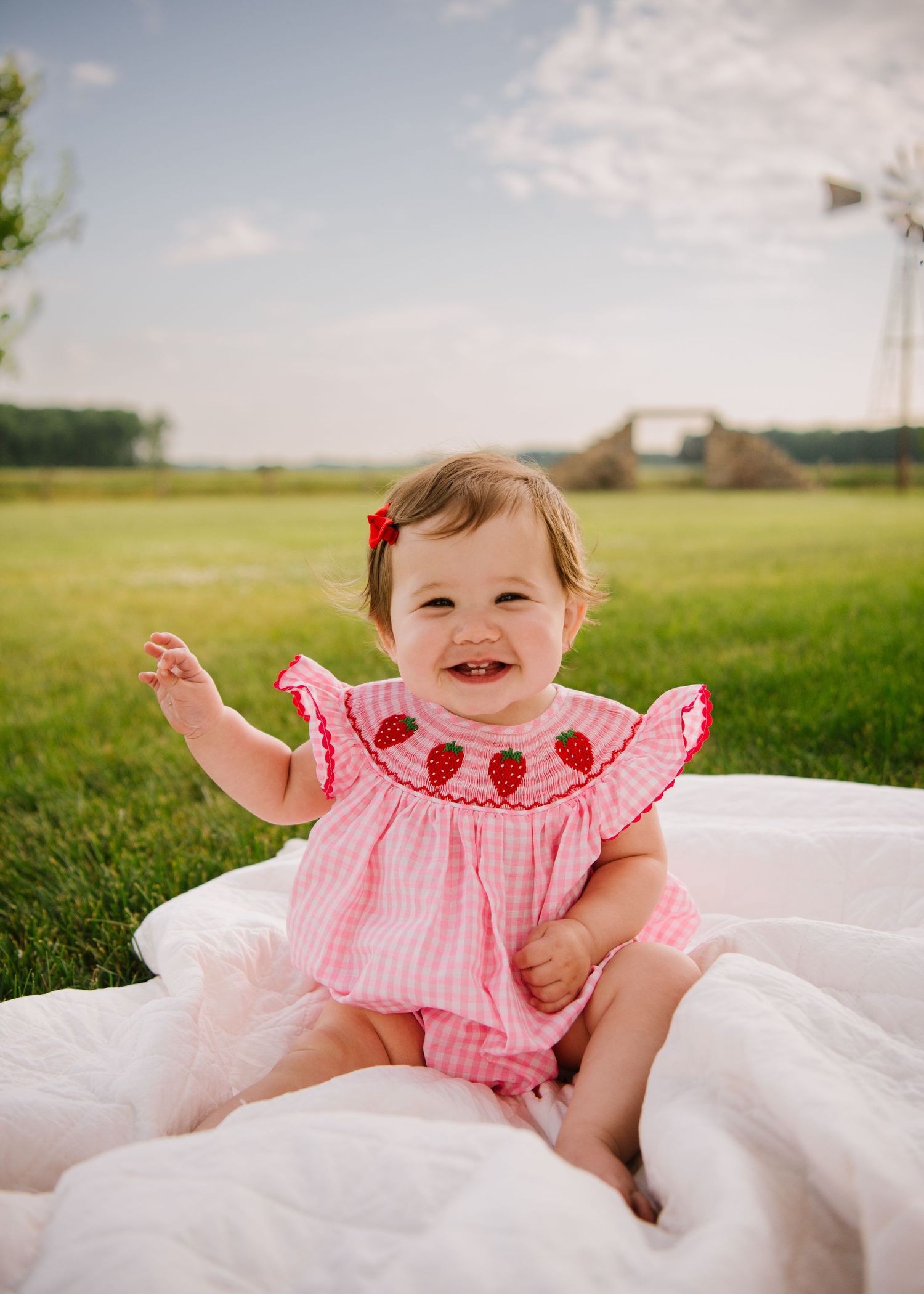 A small baby wearing a pink ruffled top giggles while laying on a white blanket in a grassy outdoor setting.