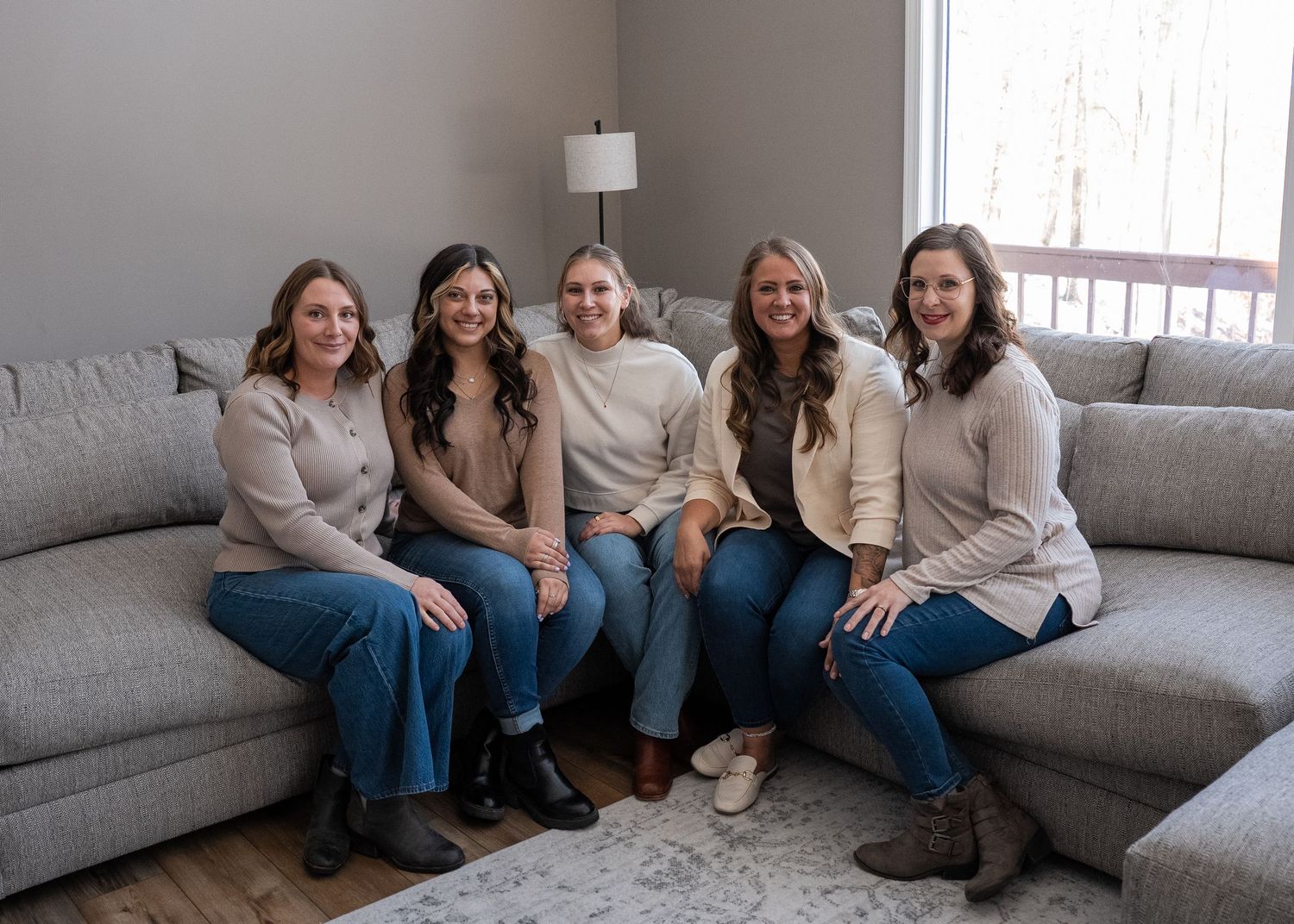 Five women smiling together on a gray sectional sofa in a modern living room setting.