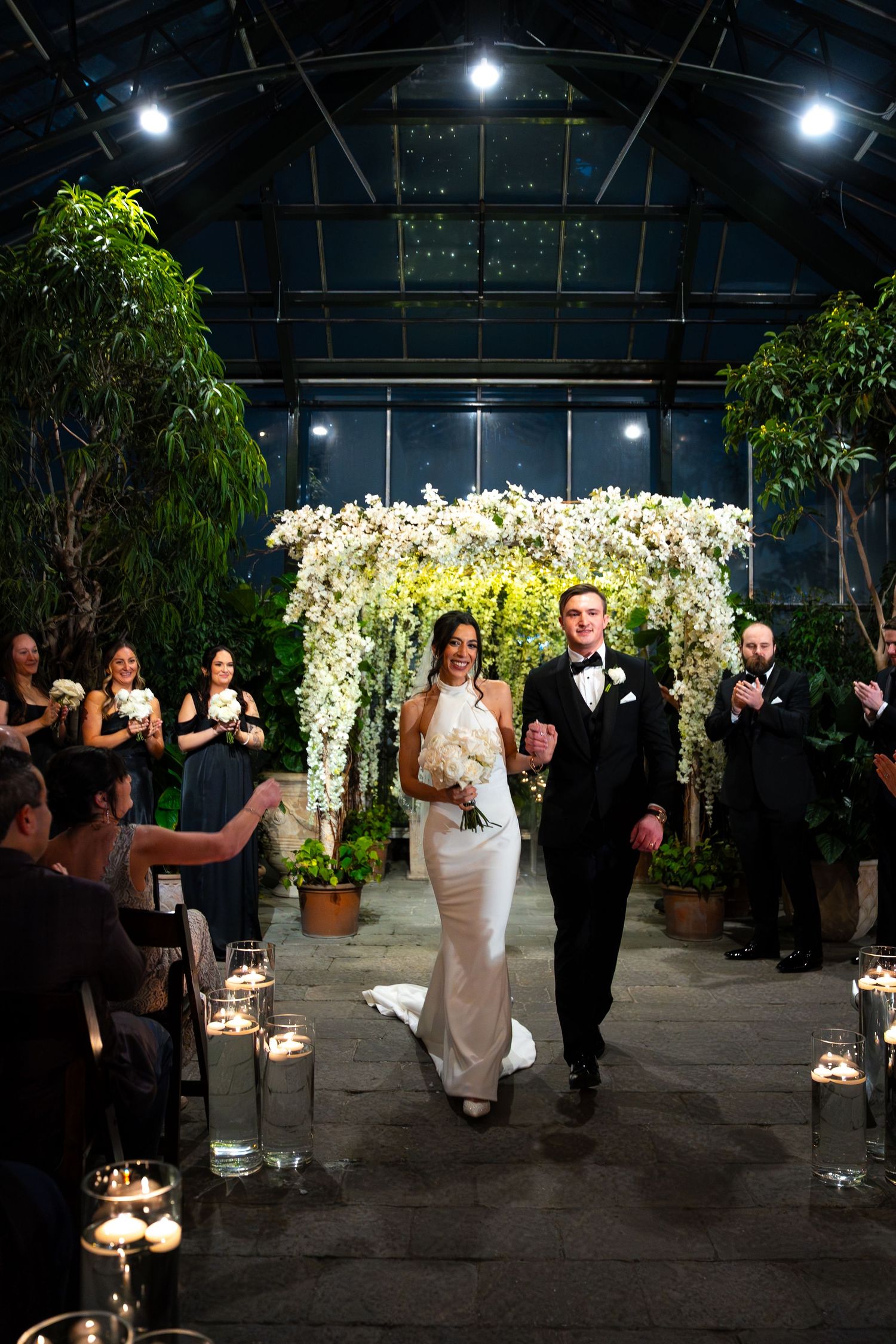 Bride and groom walk down aisle in elegant greenhouse wedding venue with floral arch and candlelit path.