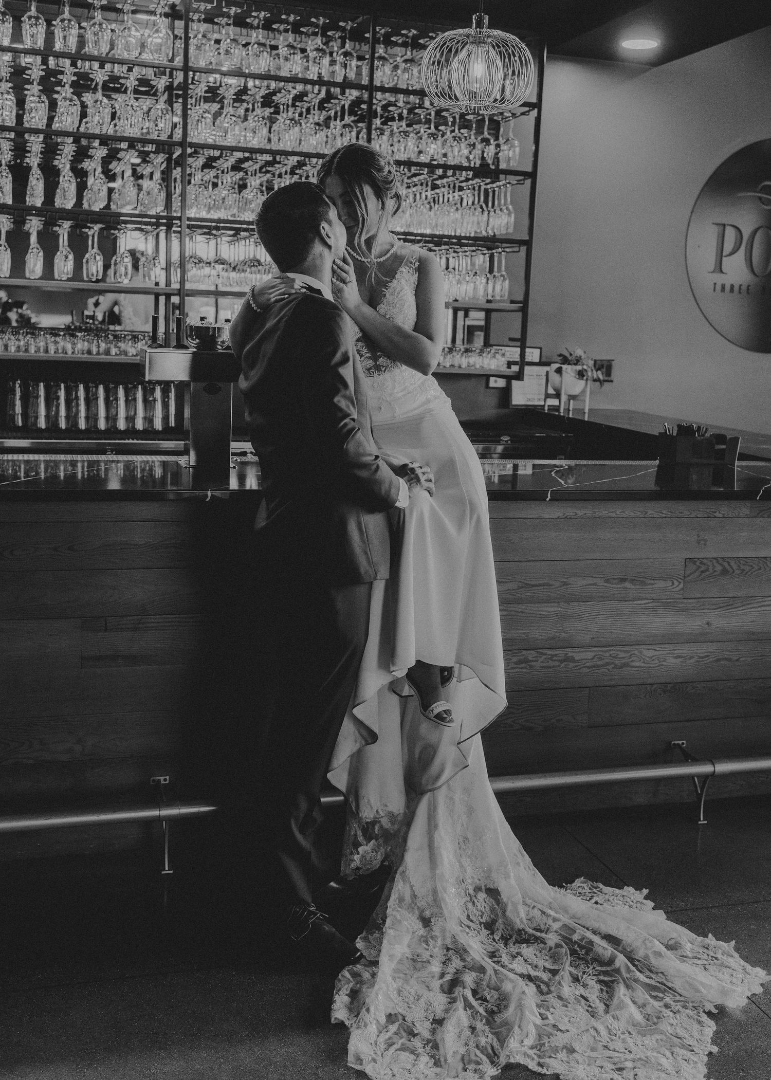 A romantic black and white wedding scene showing a couple dancing in a bar with backlit glass shelving.