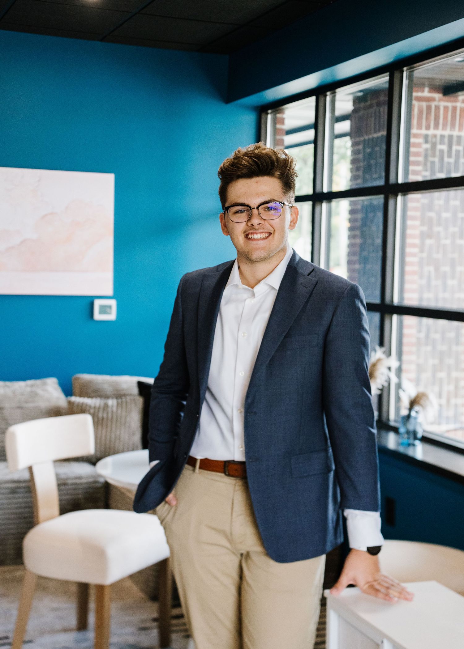 Professional wearing a navy blazer and khaki pants stands confidently in a modern office space with blue walls.
