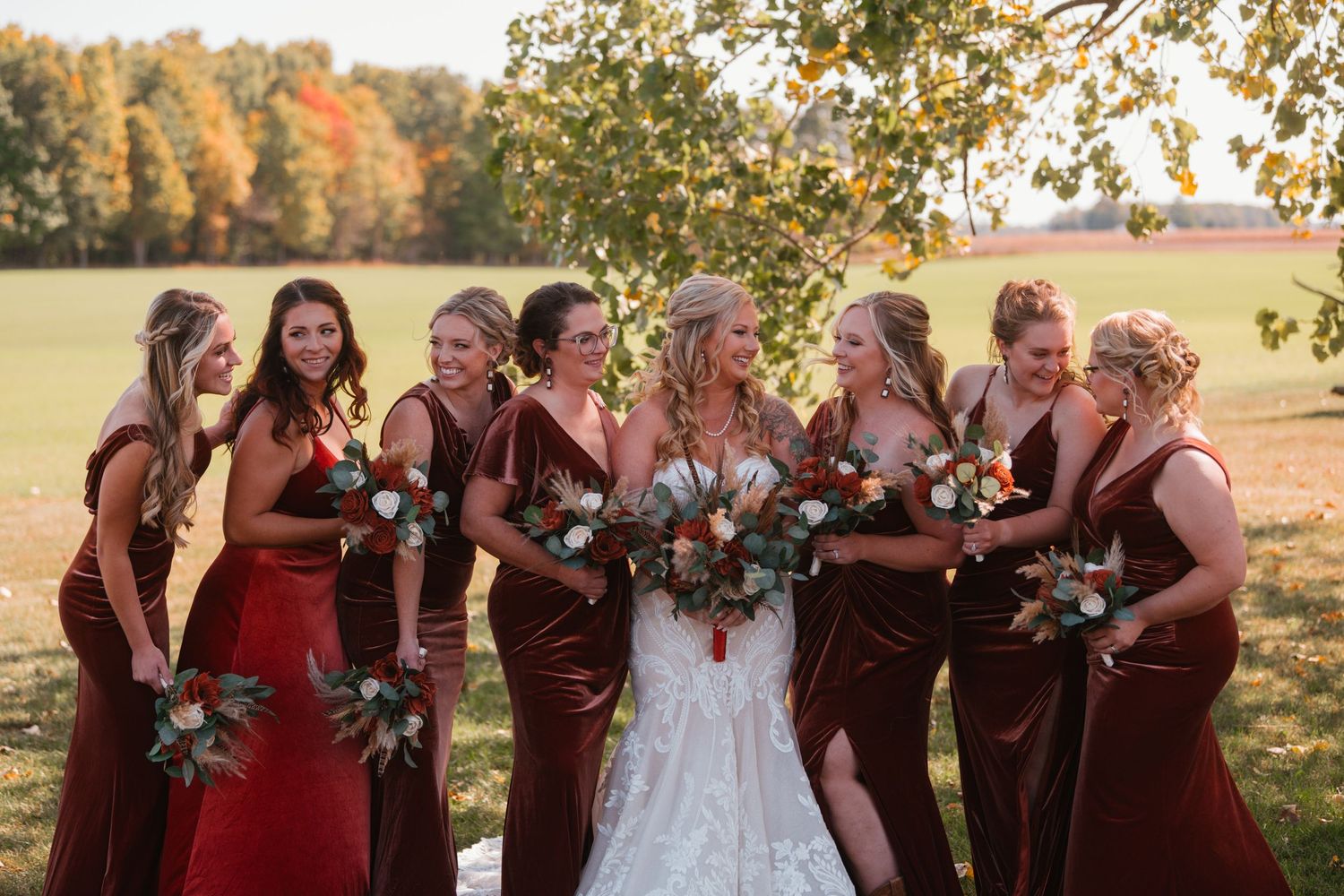 A wedding party in burgundy bridesmaids dresses and white gown gather outdoors during golden autumn light.