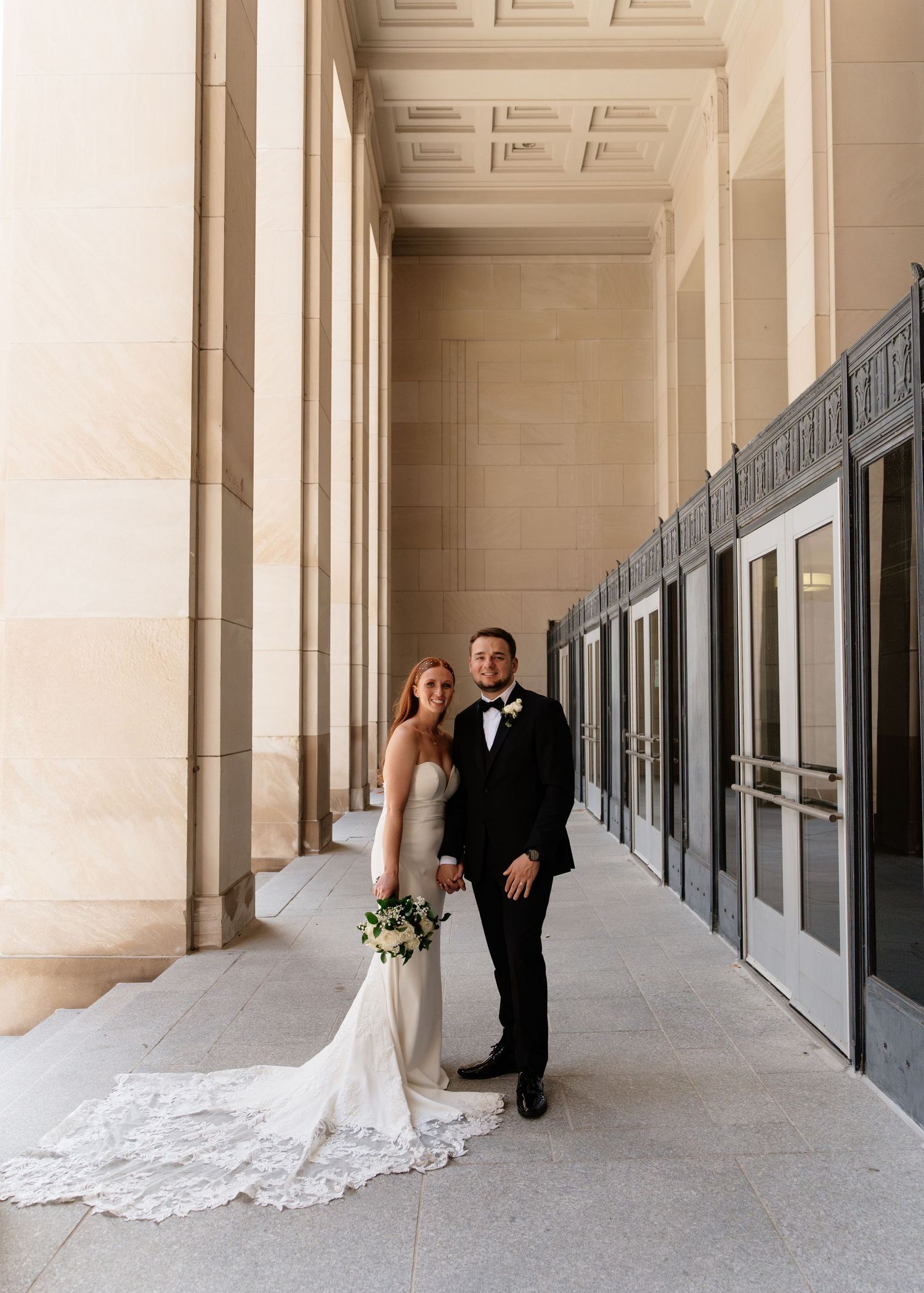 Bride in lace gown and groom in black tuxedo pose in elegant columned hallway on their wedding day.