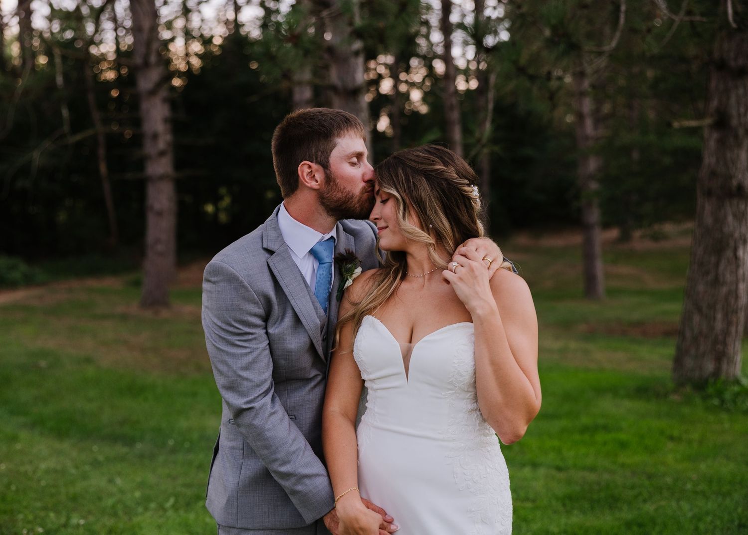 A couple shares a romantic kiss outdoors, bride in white gown and groom in gray suit, with lush green trees behind.