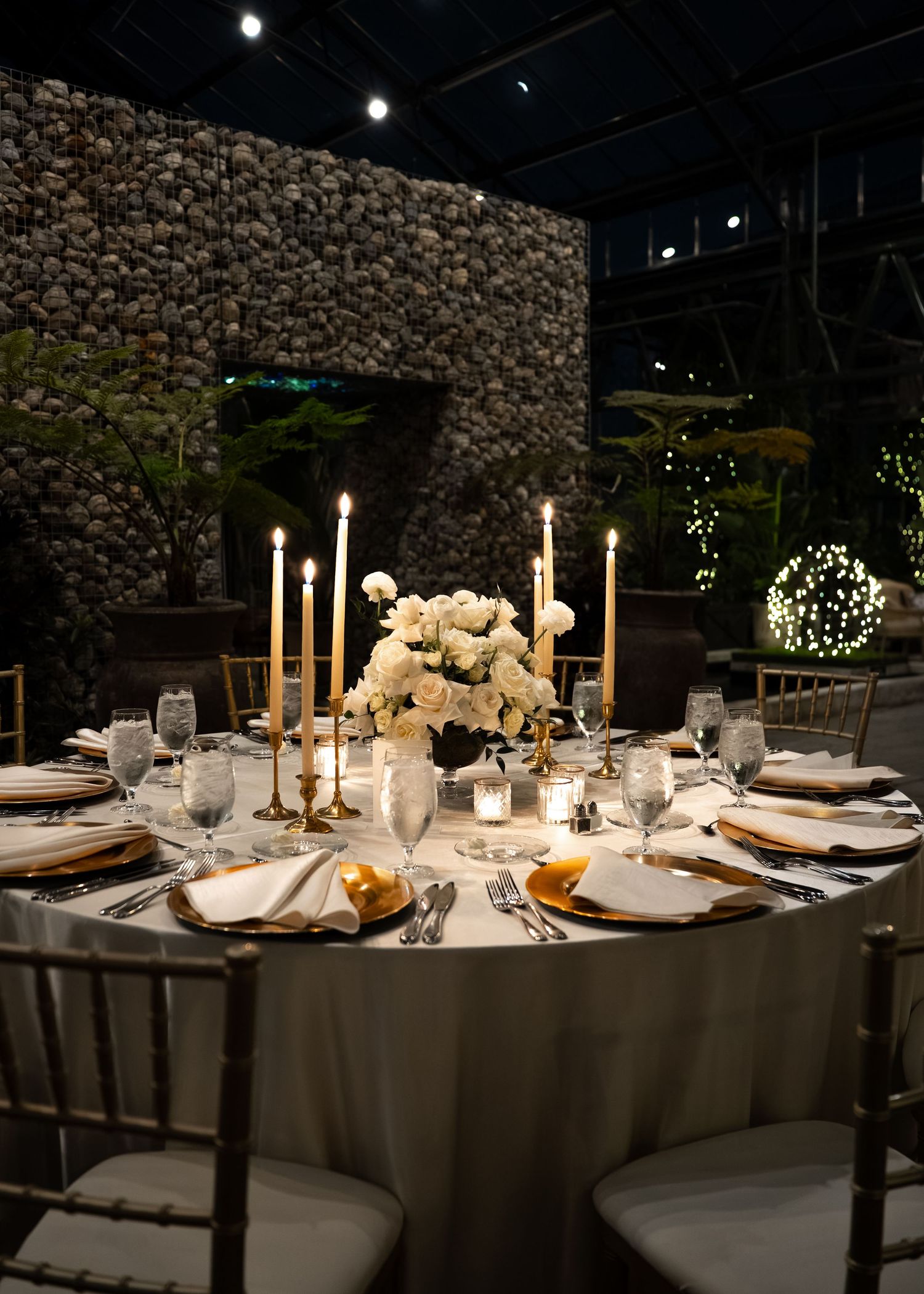 Elegant round dining table with gold candelabras, white floral centerpiece, and gold chargers in a dimly lit venue.