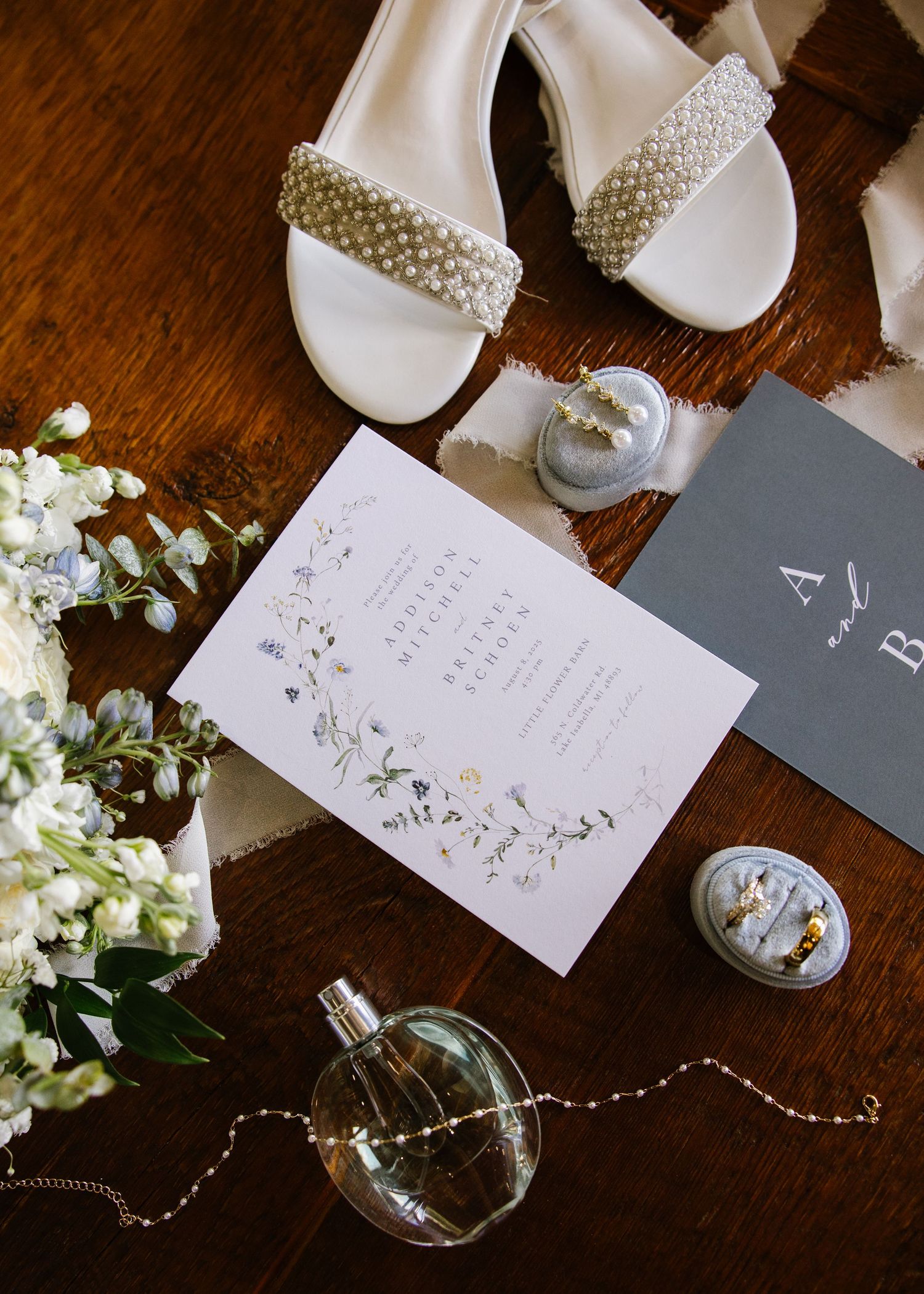 Elegant wedding flat lay with pearl heels, floral invitation, perfume, ring boxes, and white flowers on wood.