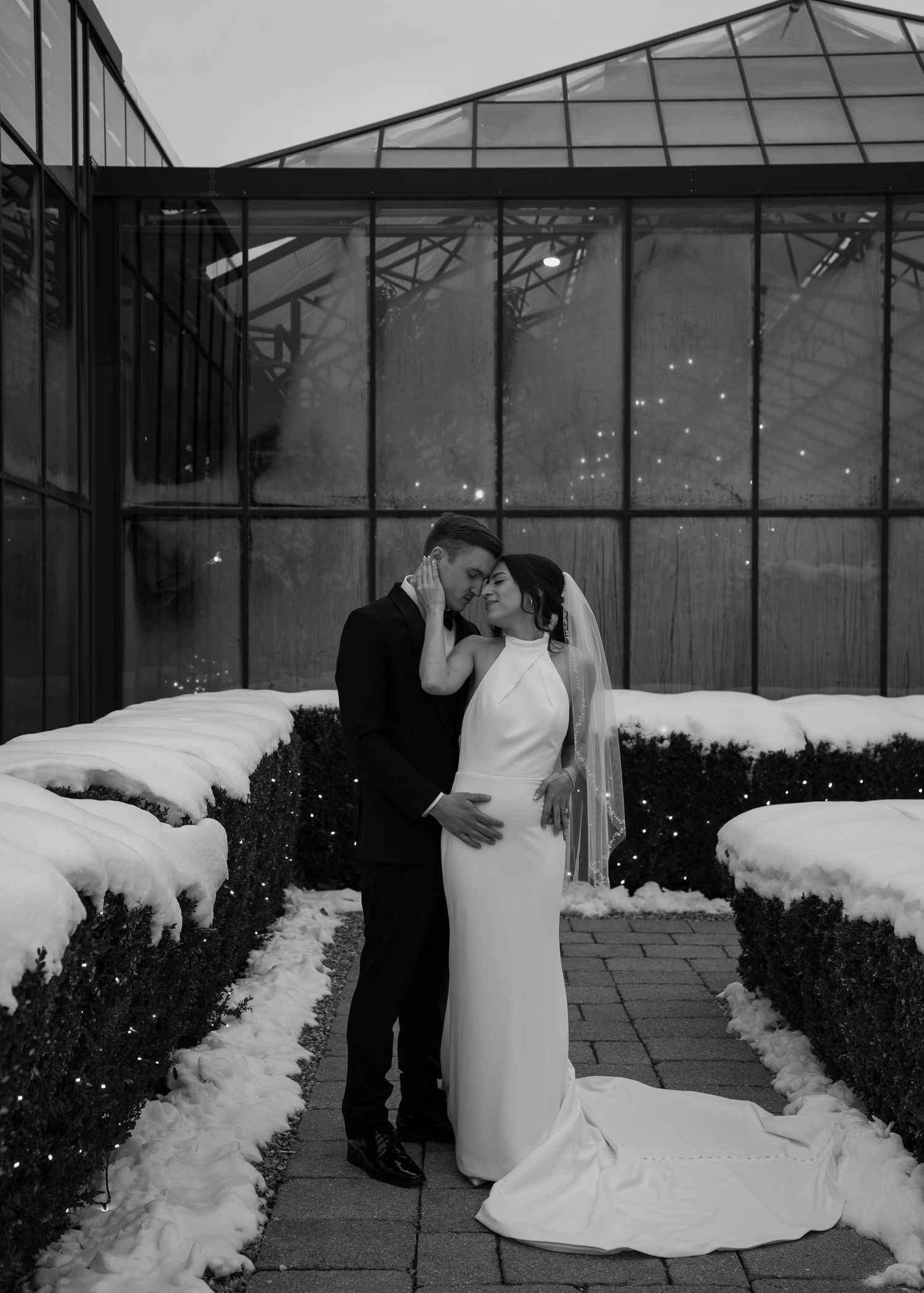 Black and white winter wedding photo of couple kissing in snowy garden outside greenhouse venue.