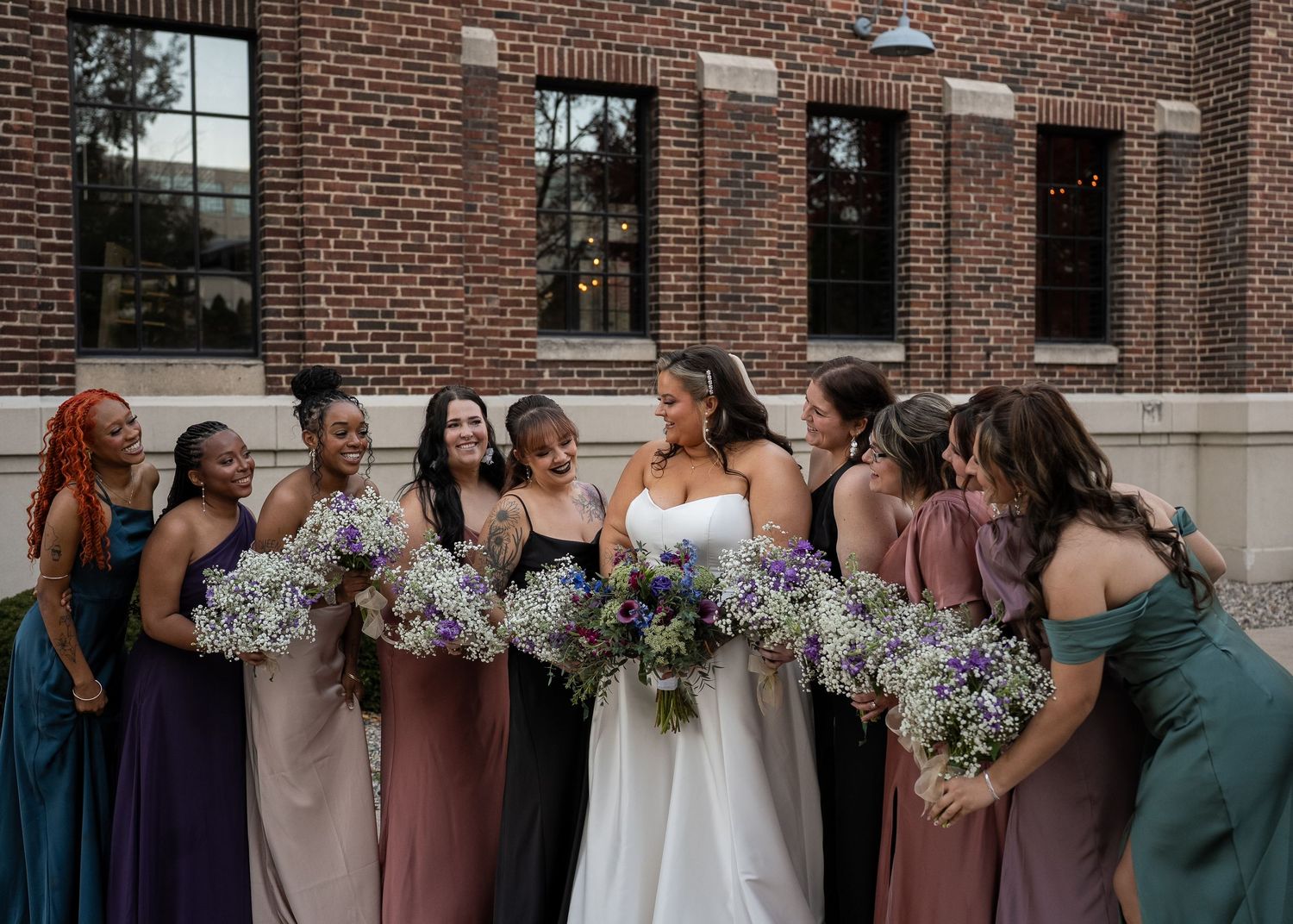 Bride in white gown stands with bridesmaids in colorful mismatched dresses holding purple and white bouquets outside brick building.