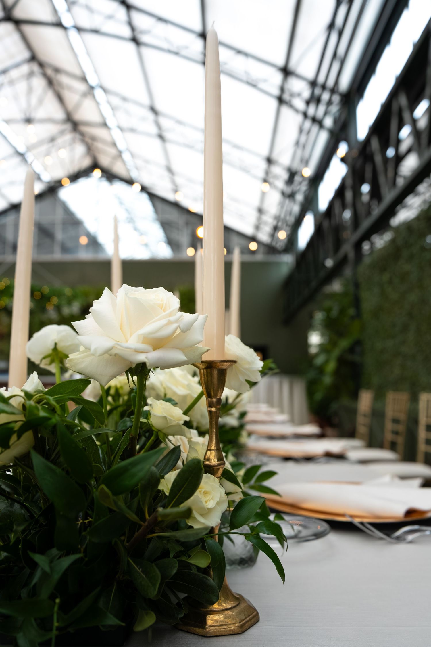 White roses and gold candelabra centerpiece on wedding table in elegant greenhouse venue with string lights.