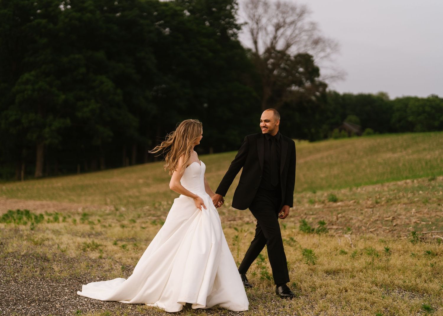 A long white wedding dress train drags gracefully across a grassy hill while walking in an outdoor field at sunset.