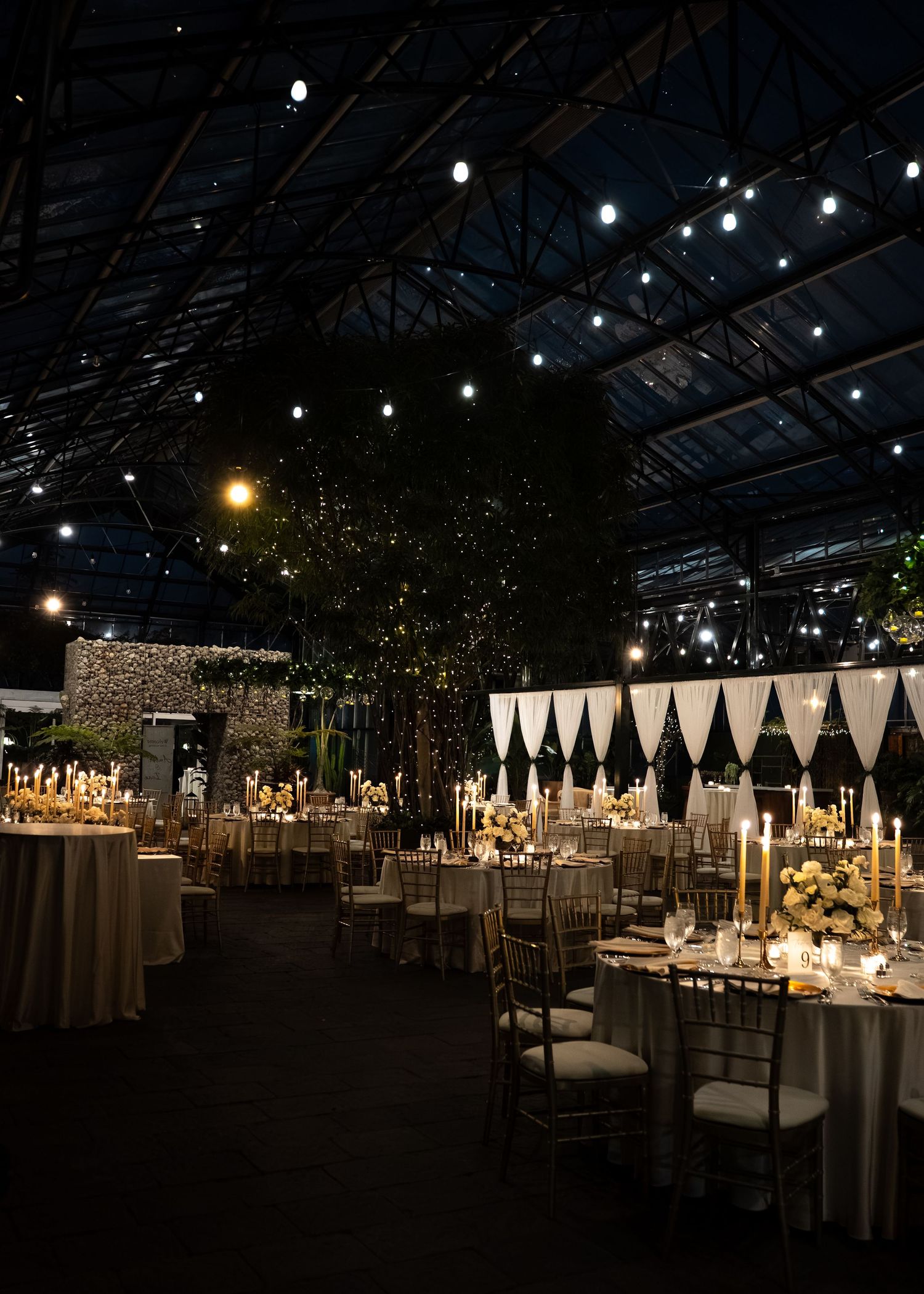 Elegant wedding reception venue with string lights, candlelit tables, floral centerpieces, and dramatic black draping at night.