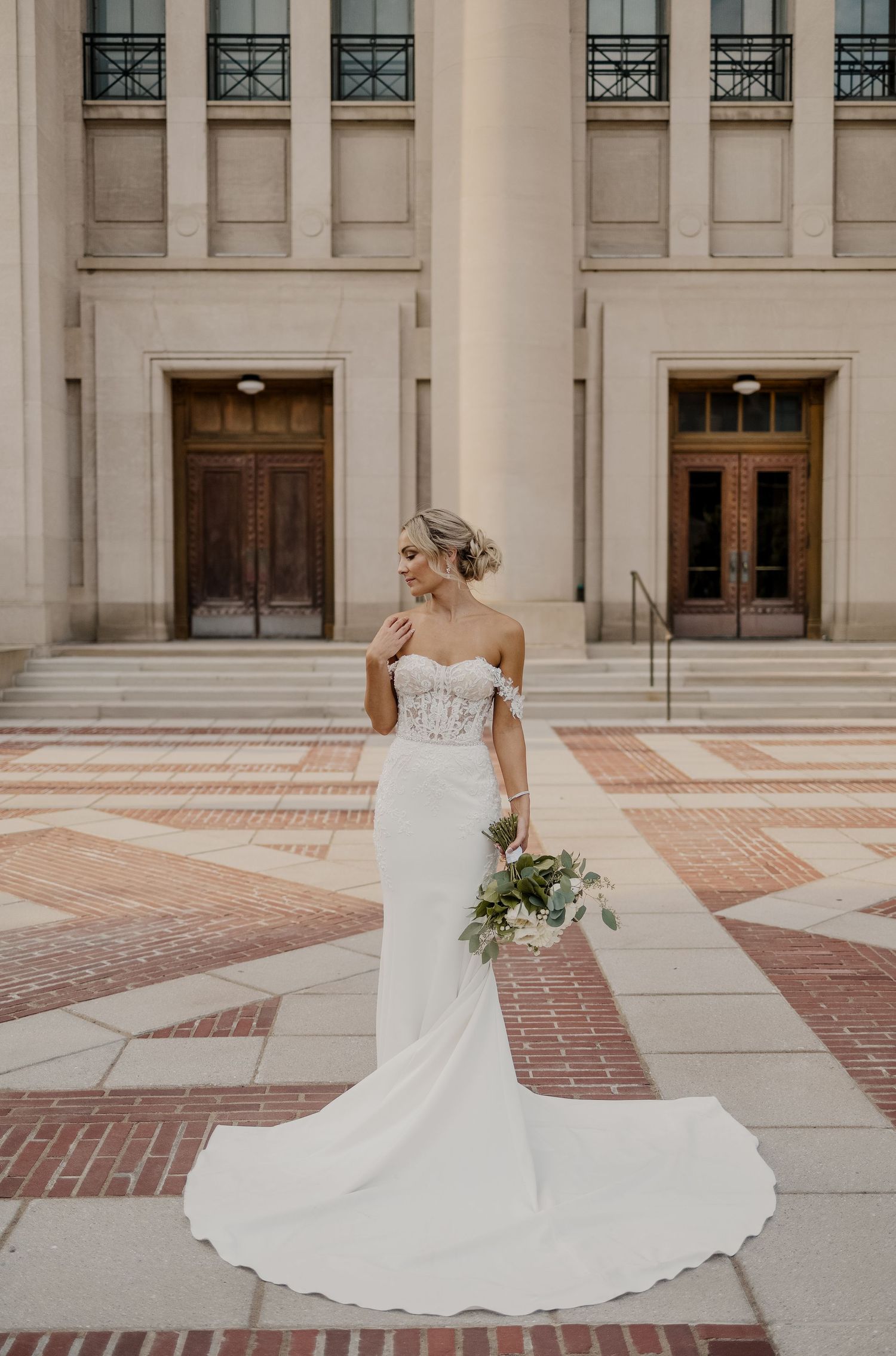Elegant bride in off-shoulder fitted white gown with train poses on ornate brick and stone courtyard near classical building.