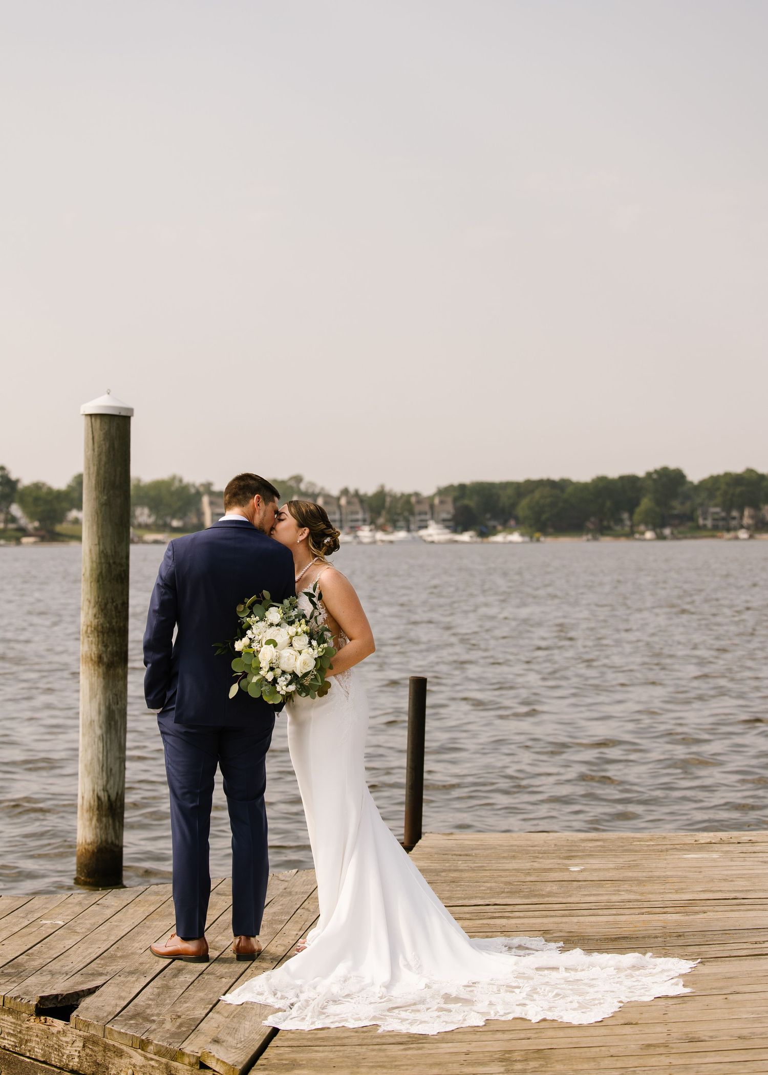 A couple shares an intimate moment on a waterfront dock with white floral bouquet and flowing wedding dress train.