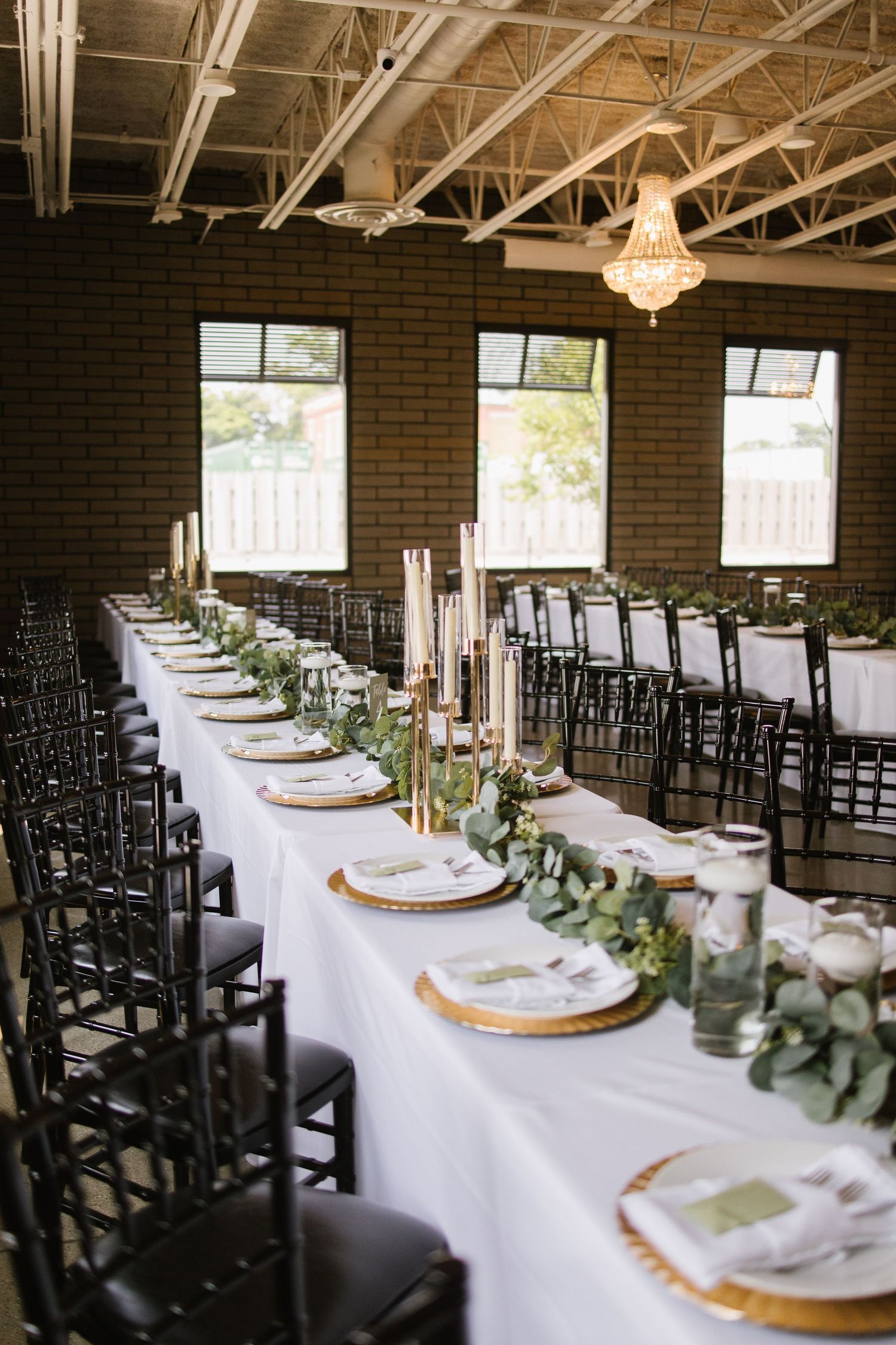 Elegant wedding reception table with white linens, gold charger plates and greenery garland set against industrial brick walls.