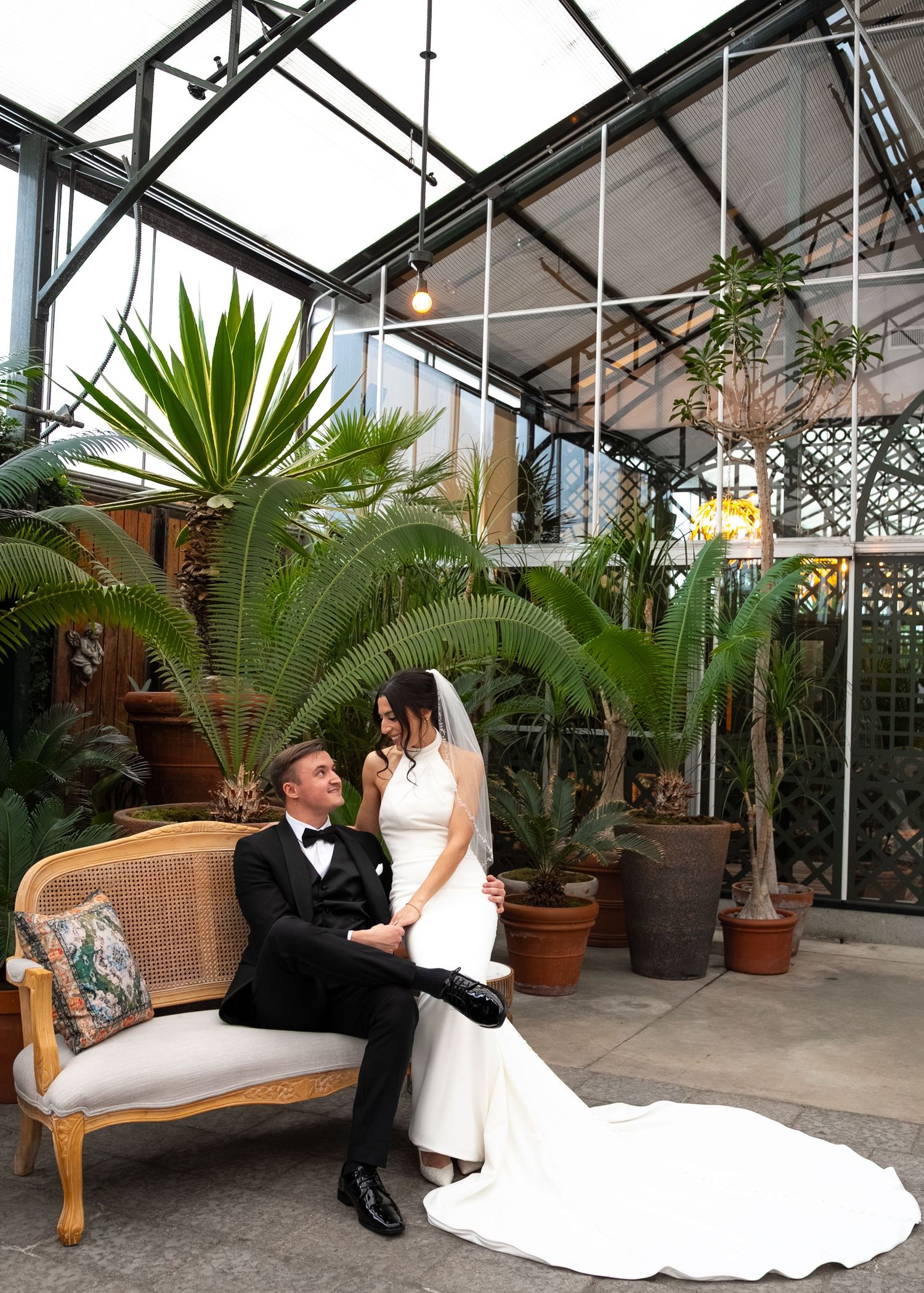 Bride and groom sitting on elegant sofa in lush greenhouse venue, surrounded by tropical plants in wedding attire.