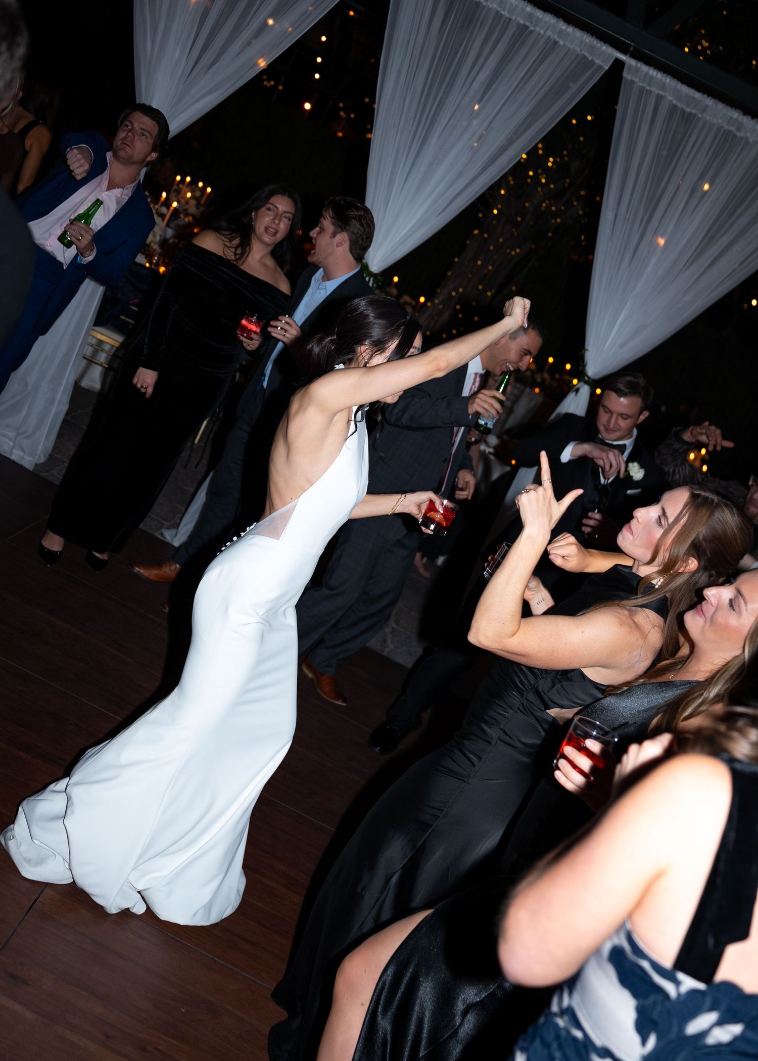 Bride in white gown dancing with guests at elegant wedding reception with draped white curtains and festive lighting.