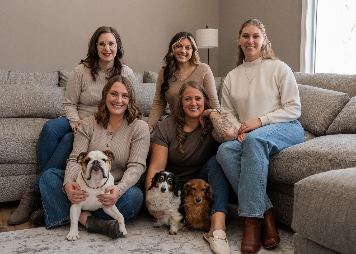 Five women smiling together on a couch with two dogs, a bulldog and a spaniel, in a cozy living room.