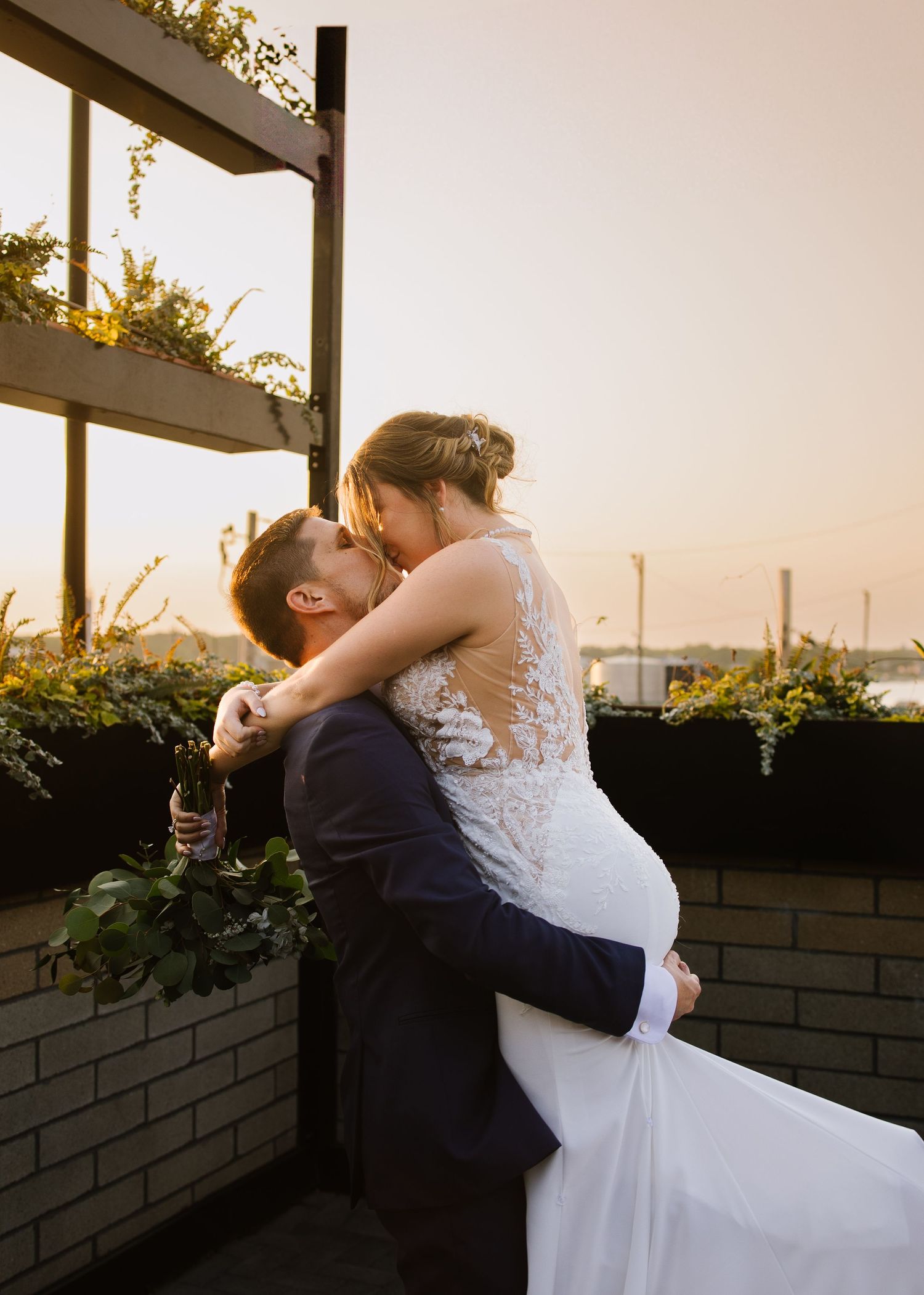A romantic wedding couple embraces during sunset on a rooftop terrace with greenery and city views in the background.