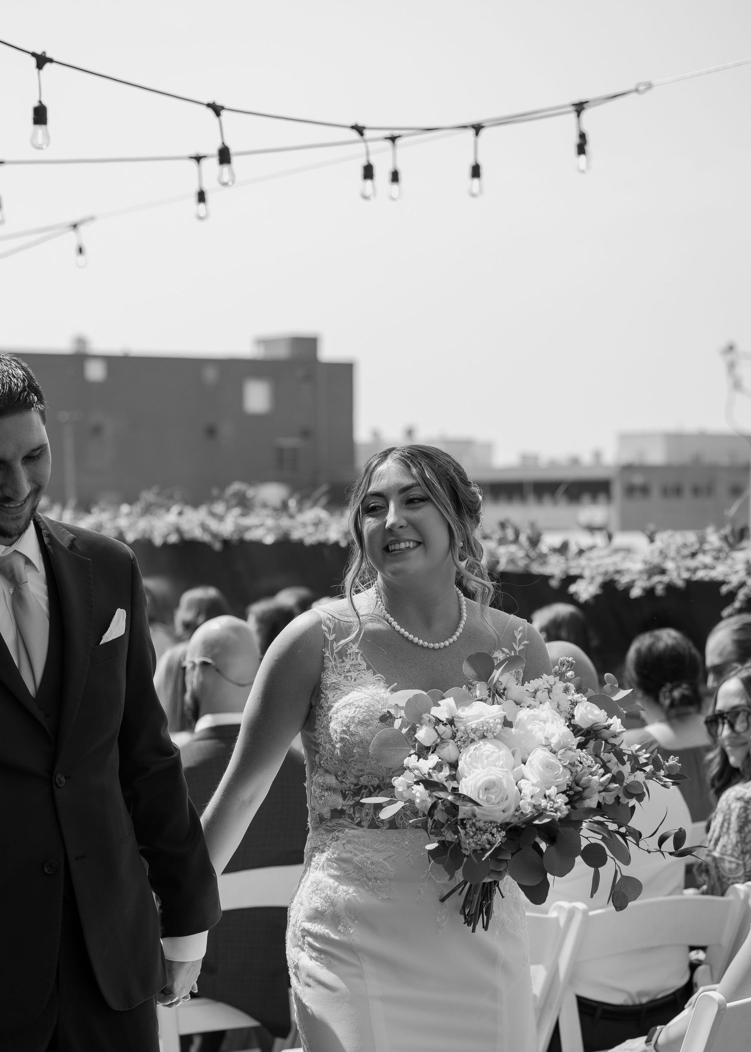 Outdoor wedding reception celebration with guests gathered under string lights at dusk in an industrial setting.
