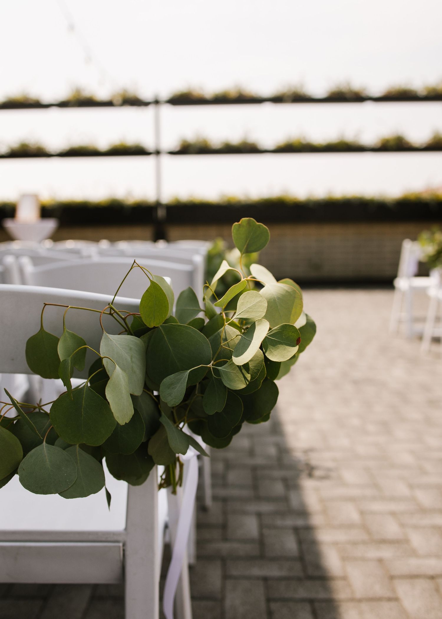 Eucalyptus leaves and greenery decorate a white folding chair at an outdoor event venue with stone pavers.