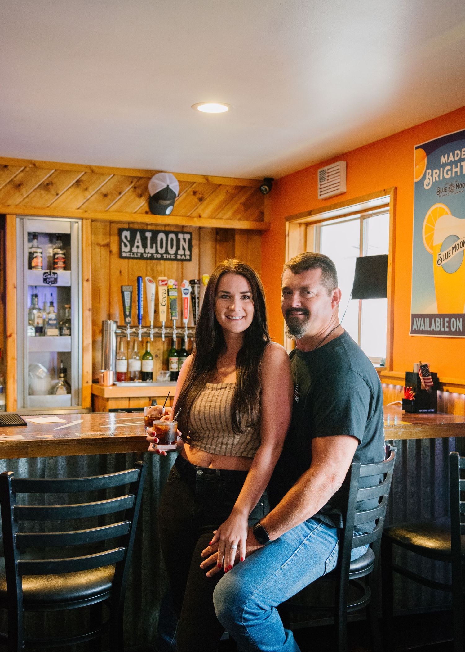 Two people pose together against an orange wall and wooden panels in a casual bar or restaurant setting.