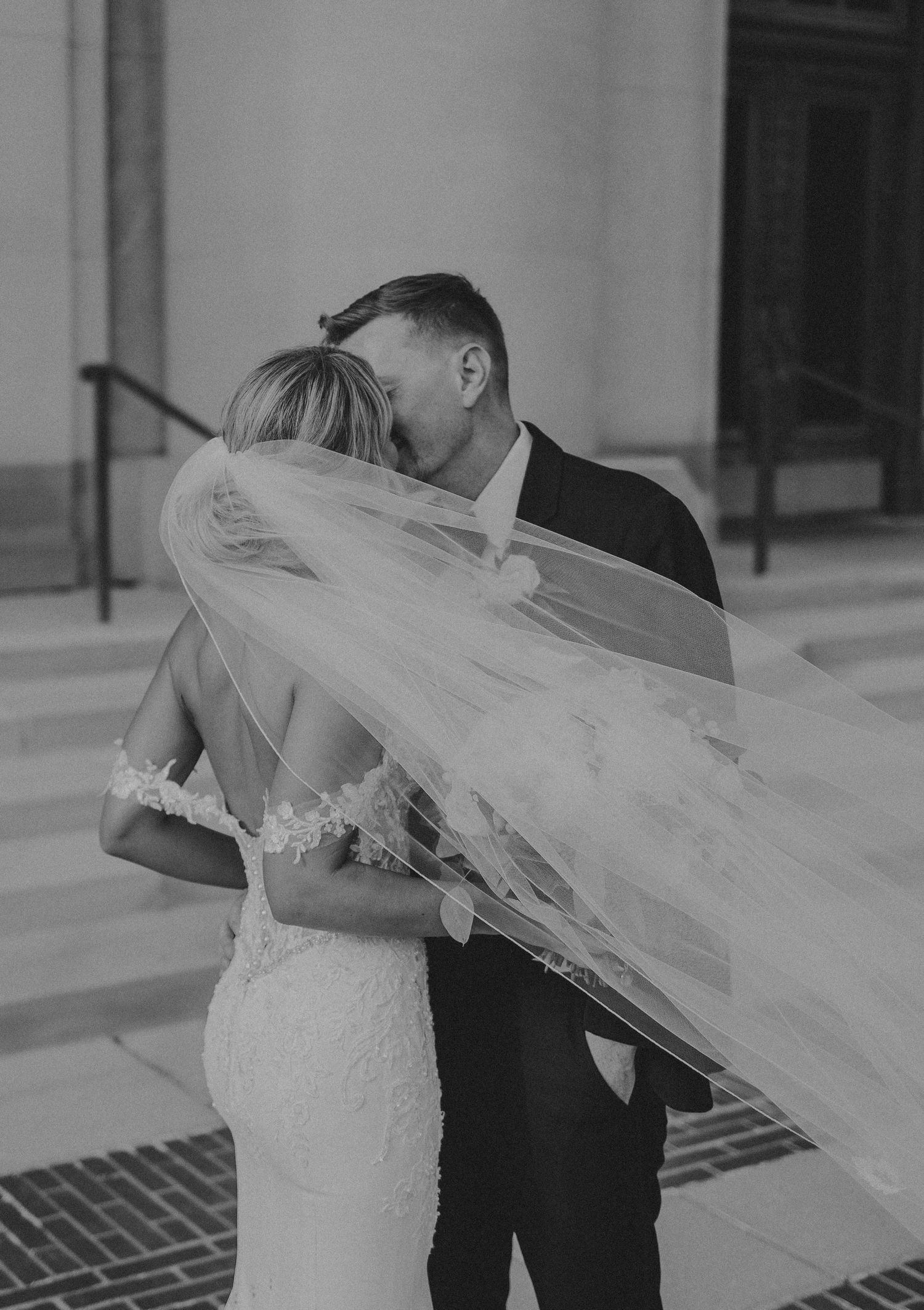 A romantic black and white wedding photo showing the couple sharing a kiss as the bride's veil flows in the breeze.