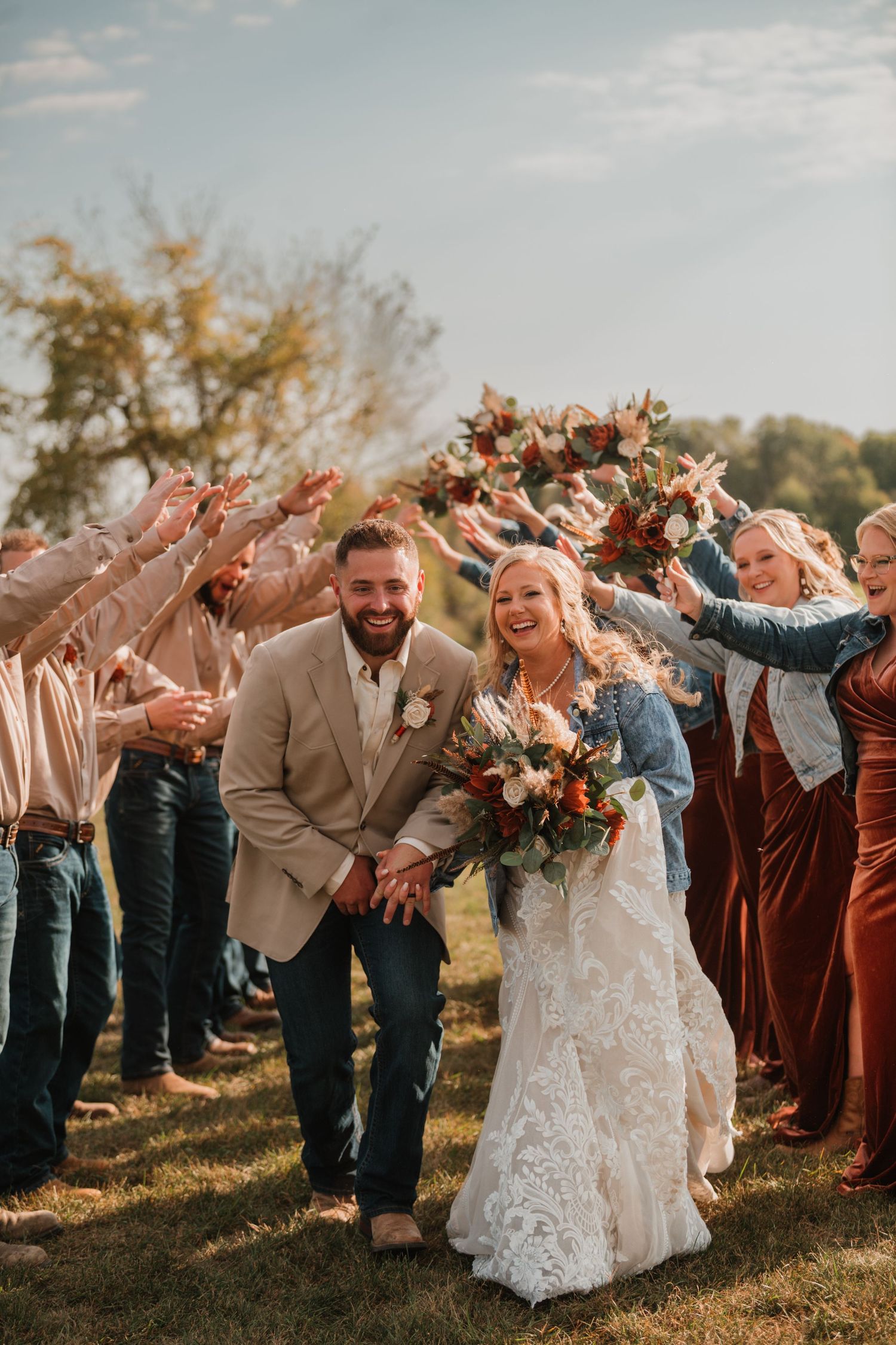 Newlyweds walk through an outdoor aisle of cheering wedding guests under golden autumn sunlight.