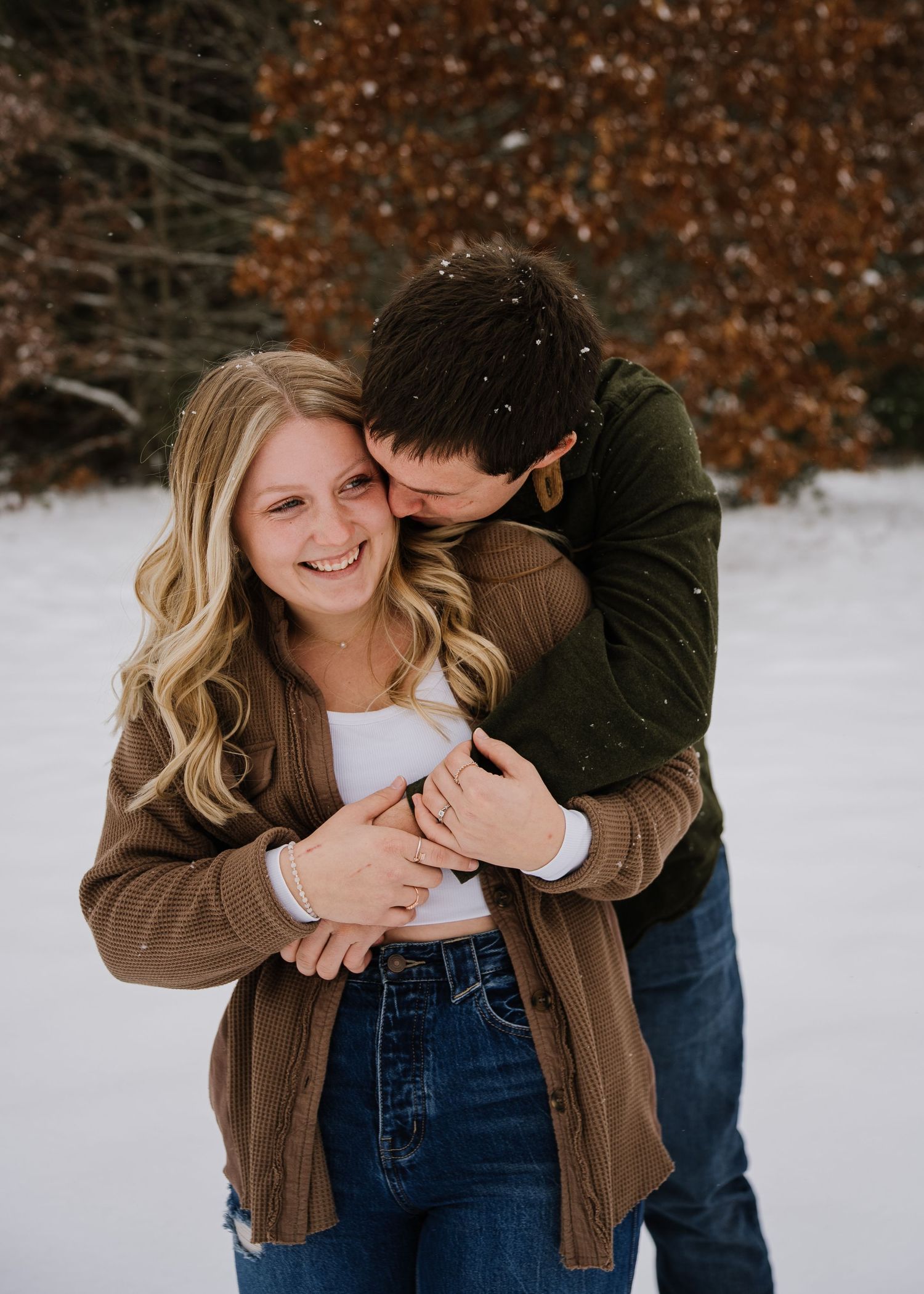 A couple embraces in a snowy winter setting with brown autumn leaves in the background.