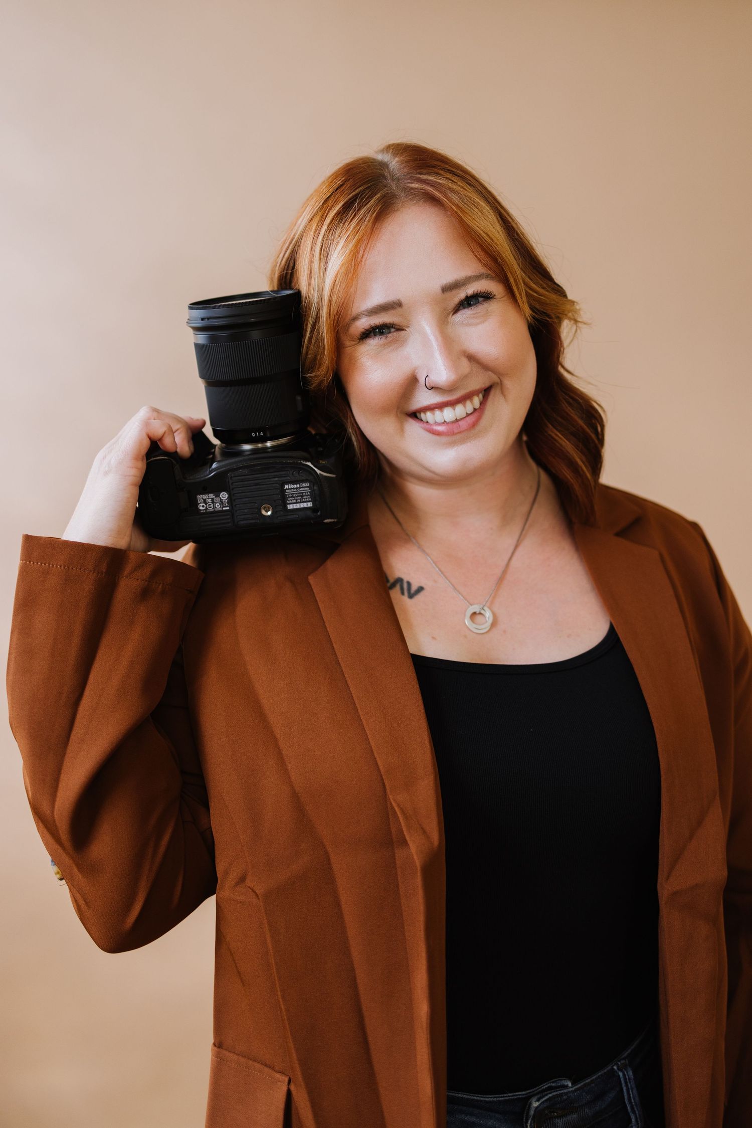 Professional photographer in brown blazer and black top poses with camera in studio against neutral background.