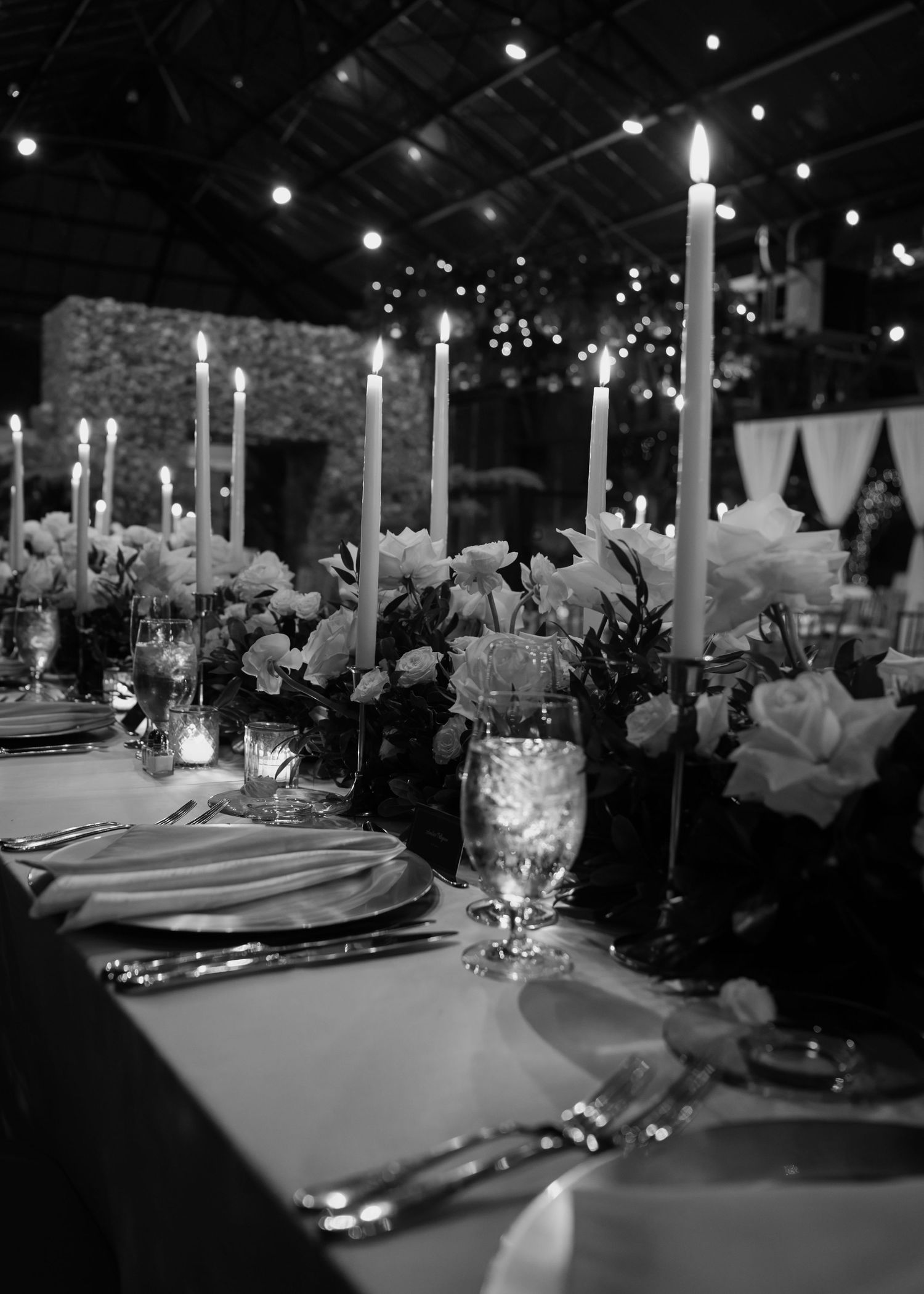 Elegant black and white wedding reception table with tall candles, floral centerpieces, and string lights in background.