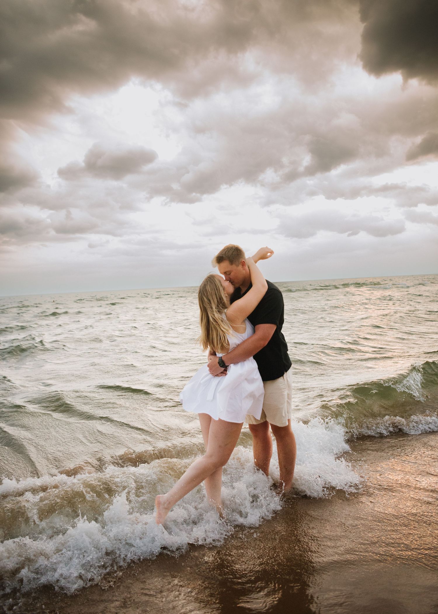 A couple embraces on a stormy beach as waves crash at their feet during sunset, creating a romantic coastal scene.