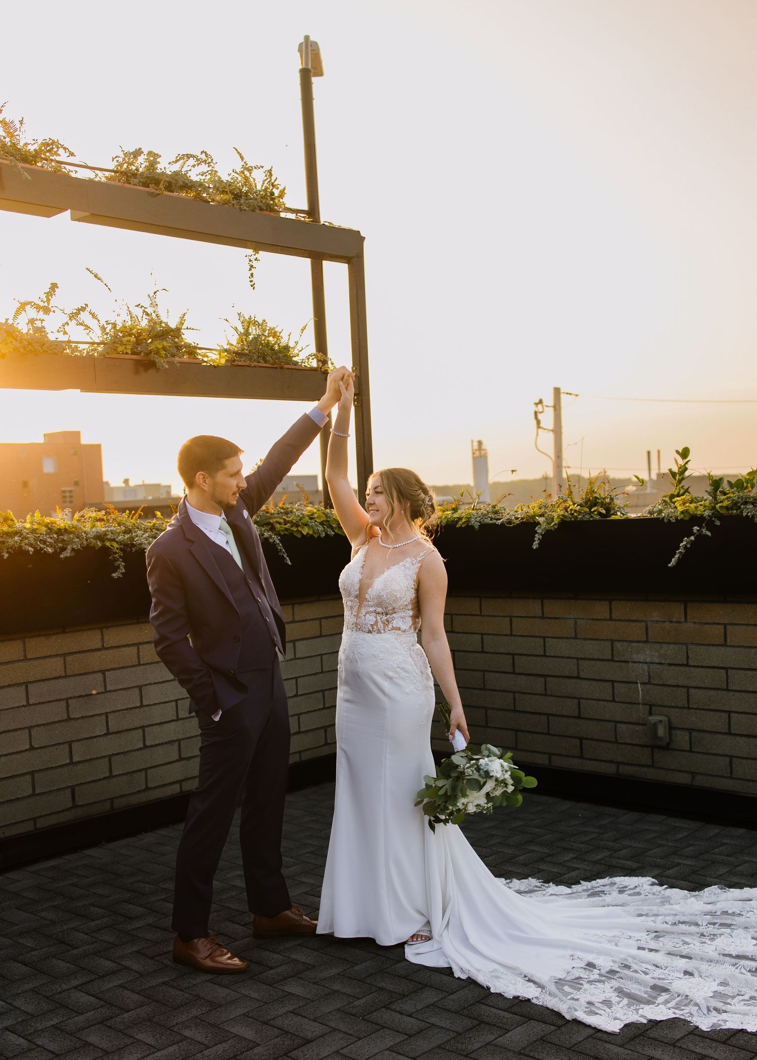 A couple in wedding attire stand on a rooftop terrace at sunset with city buildings in the background.