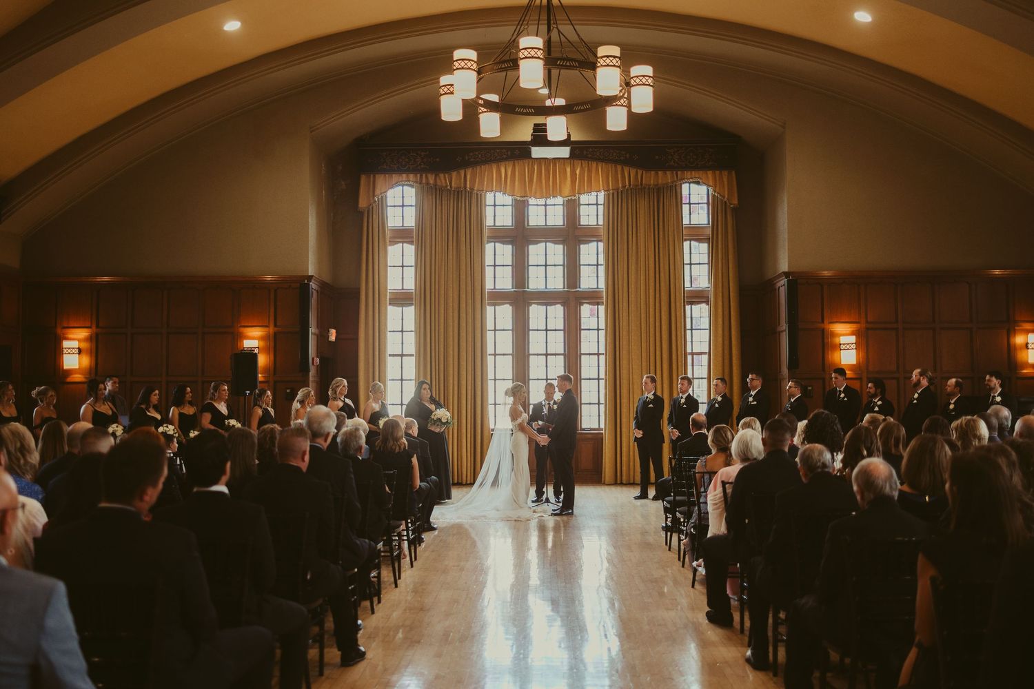 Wedding ceremony in an elegant, grand ballroom with arched windows and a chandelier while guests sit in rows of chairs.