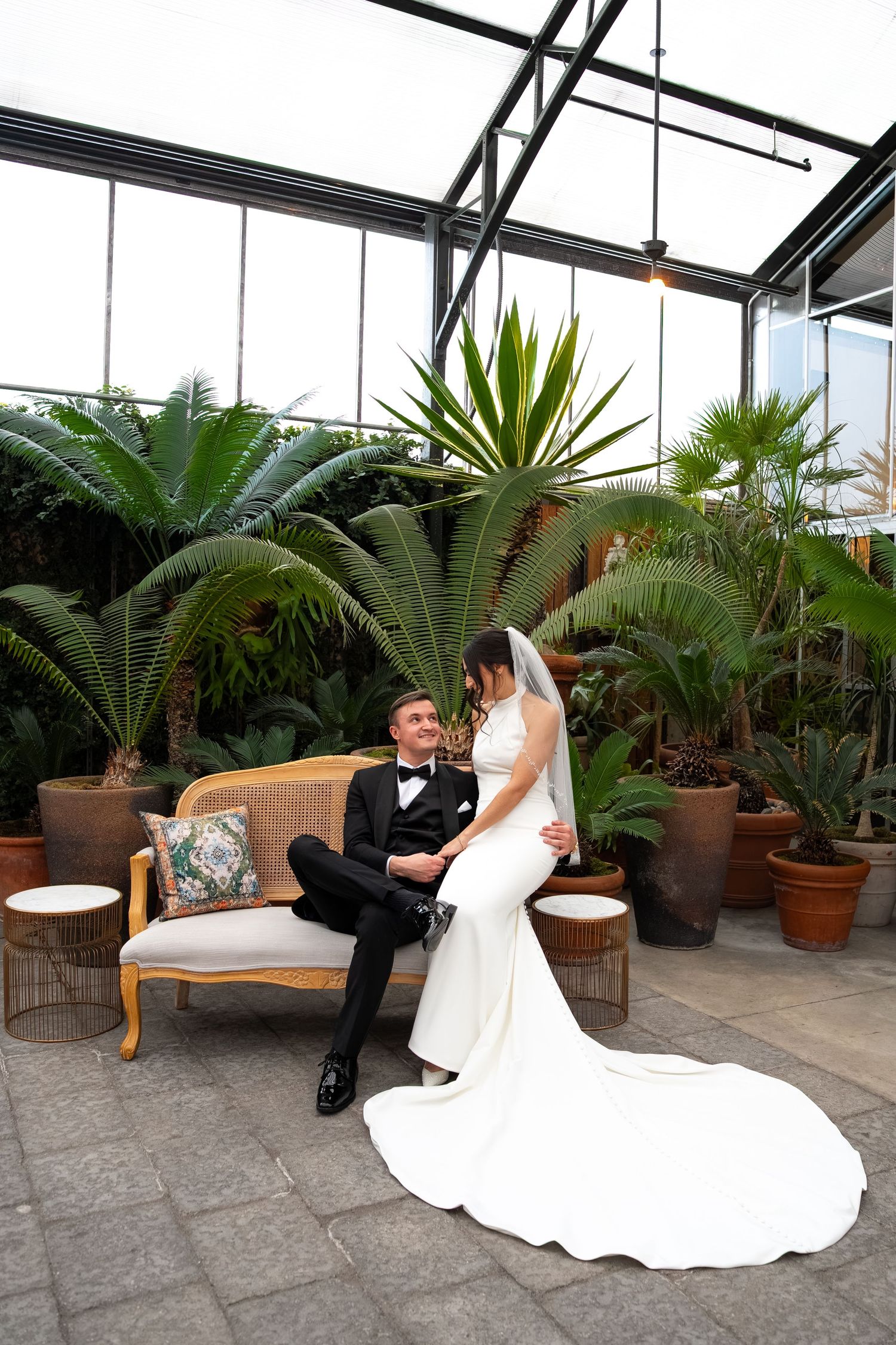 Bride and groom share a romantic moment on a vintage sofa surrounded by lush tropical plants in a glass greenhouse.