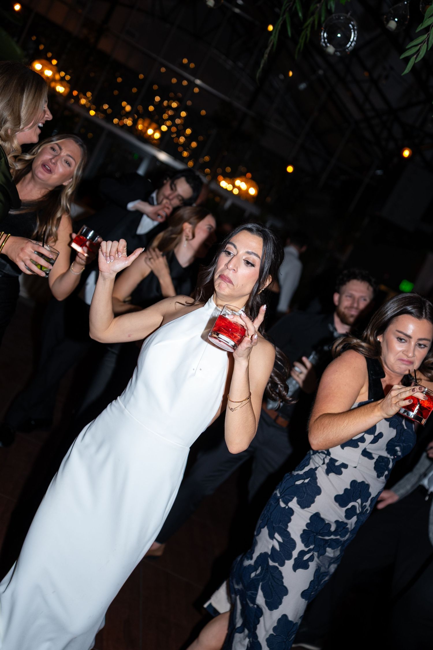 Bride in white gown dancing joyfully with guests at a wedding reception, holding a red drink.