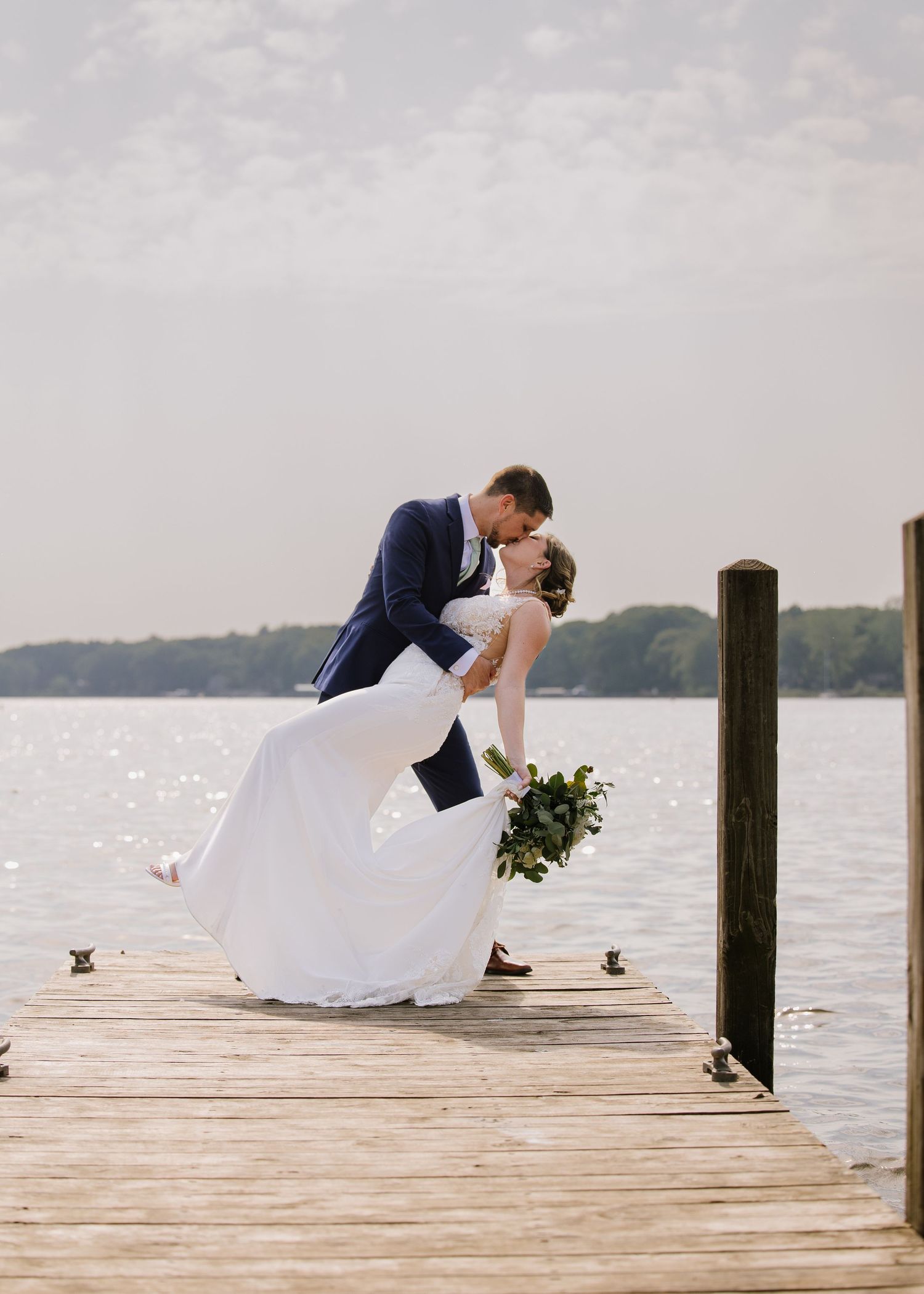 A romantic moment between newlyweds sharing a kiss on a wooden dock over a serene lake at sunset.