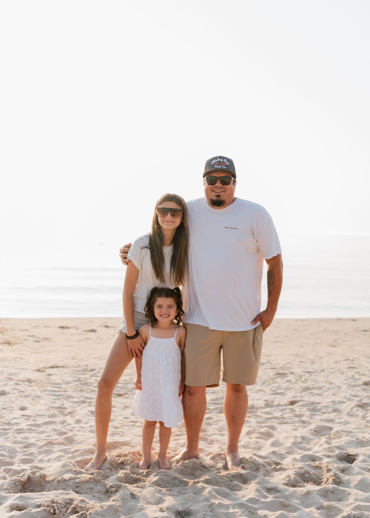 Family of three posing on sandy beach, two adults and young girl in white dress, ocean backdrop on sunny day.