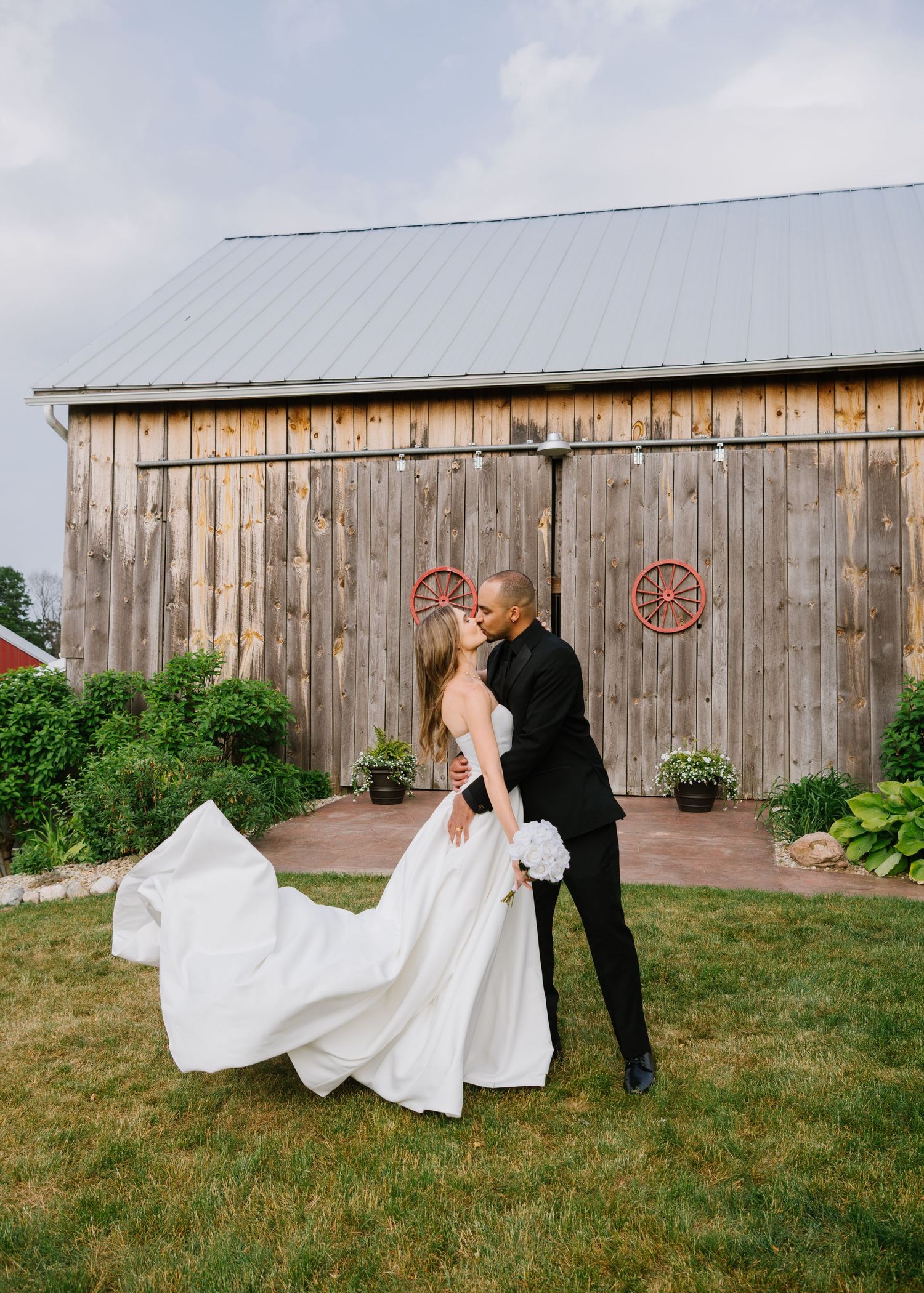 Romantic wedding couple embraces outside rustic wooden barn with flowing white dress on green lawn.