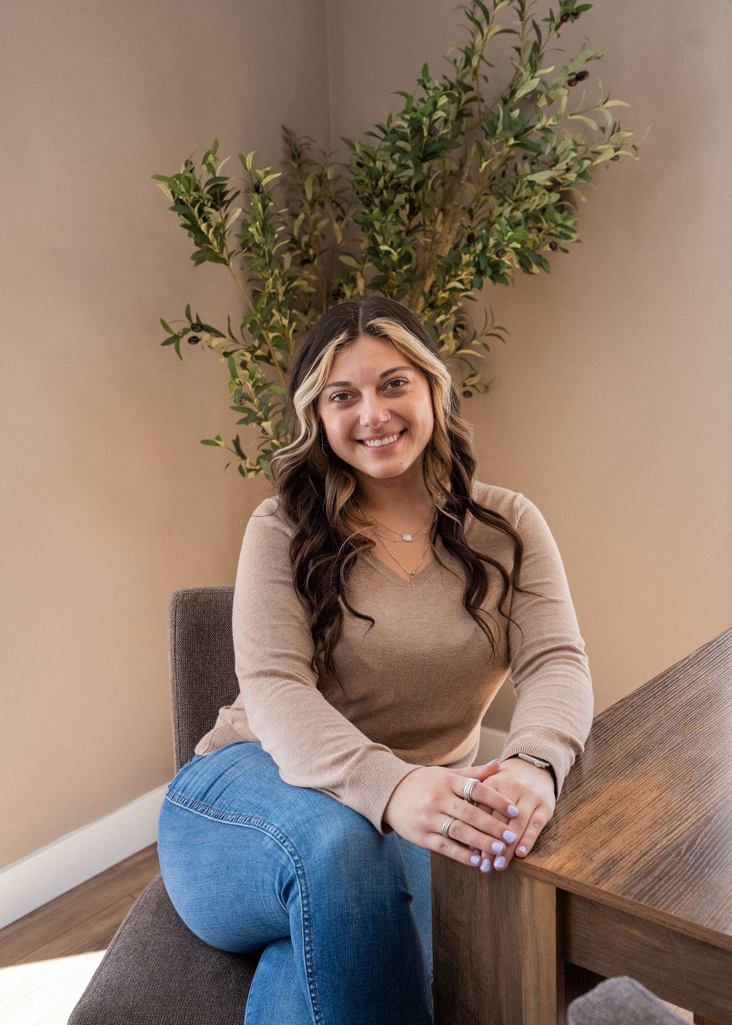 Young woman with long dark hair smiling, seated in a chair wearing a tan top and jeans, with a green plant behind her.