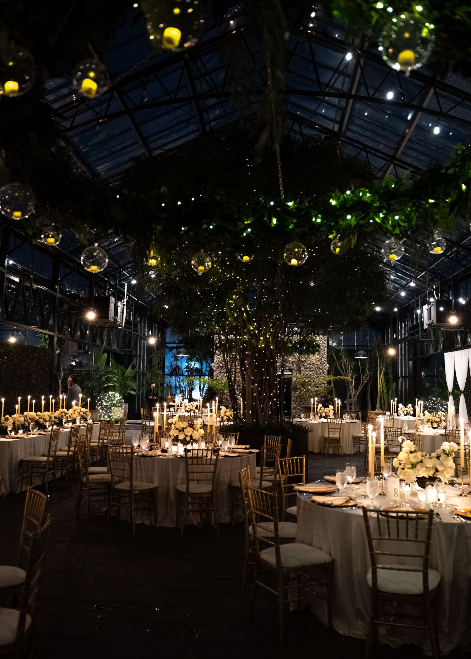 Elegant greenhouse wedding reception with gold chairs, candlelit tables, lush greenery, and hanging glass orbs under a glass ceiling.
