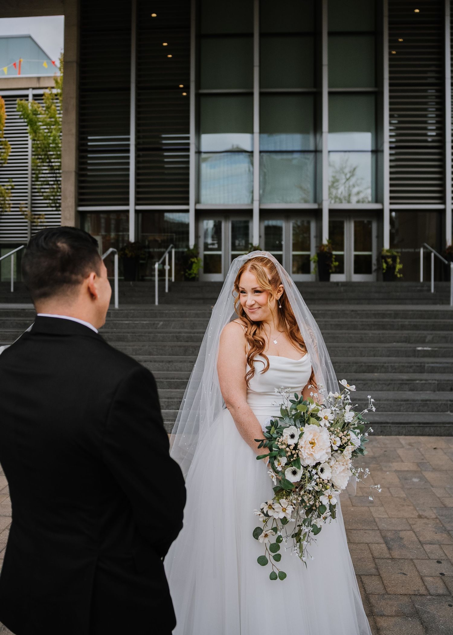 Bride in white gown and veil holds cascading bouquet, smiling at groom in black suit outside modern building.